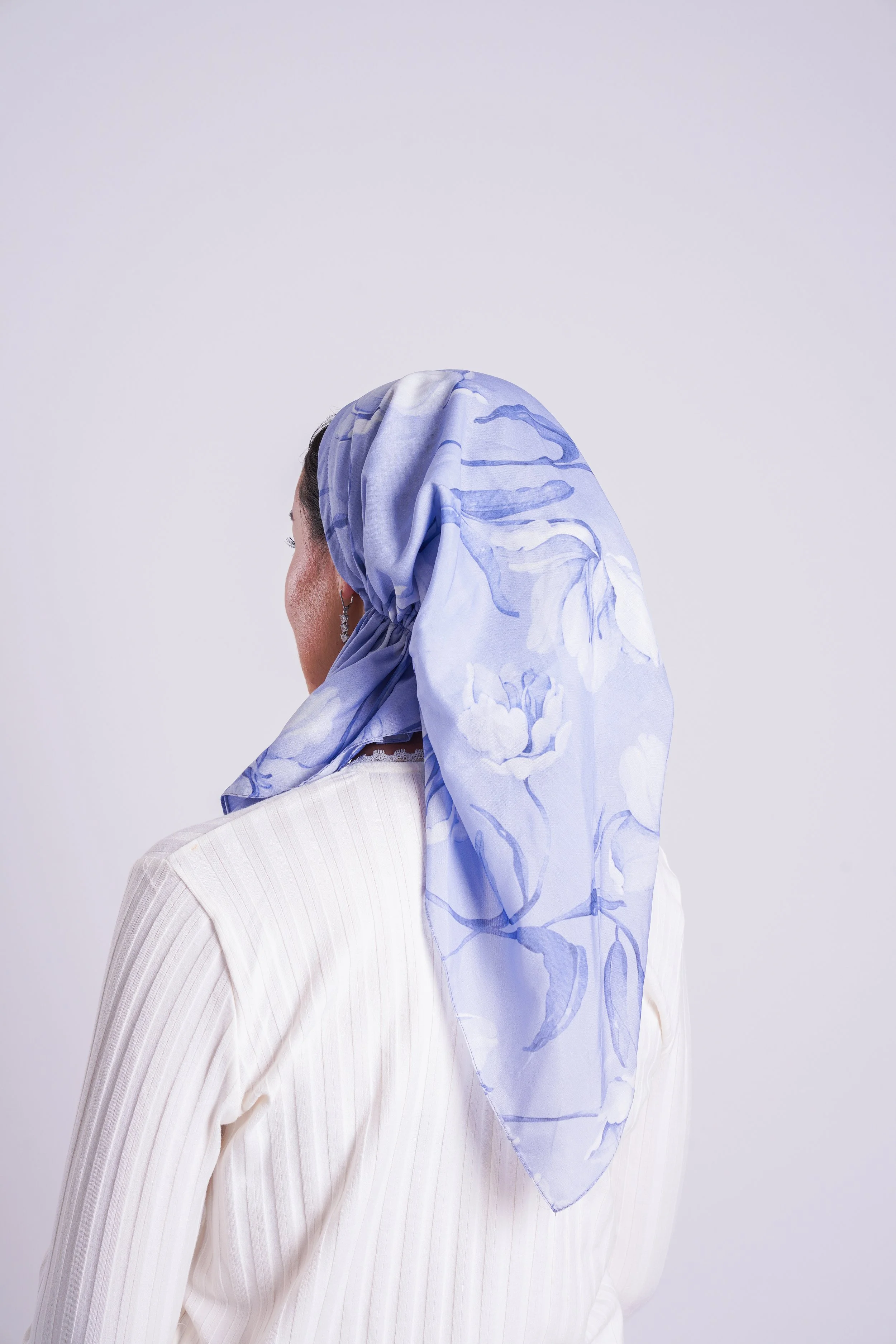 Back view of a woman wearing a light-colored, vertically striped blazer and a blue floral silk headscarf against a plain white background.