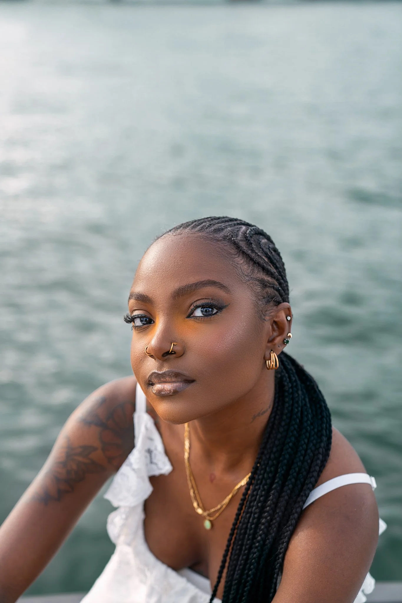 A young woman with braids, wearing jewelry, dressed in a white top, near water.