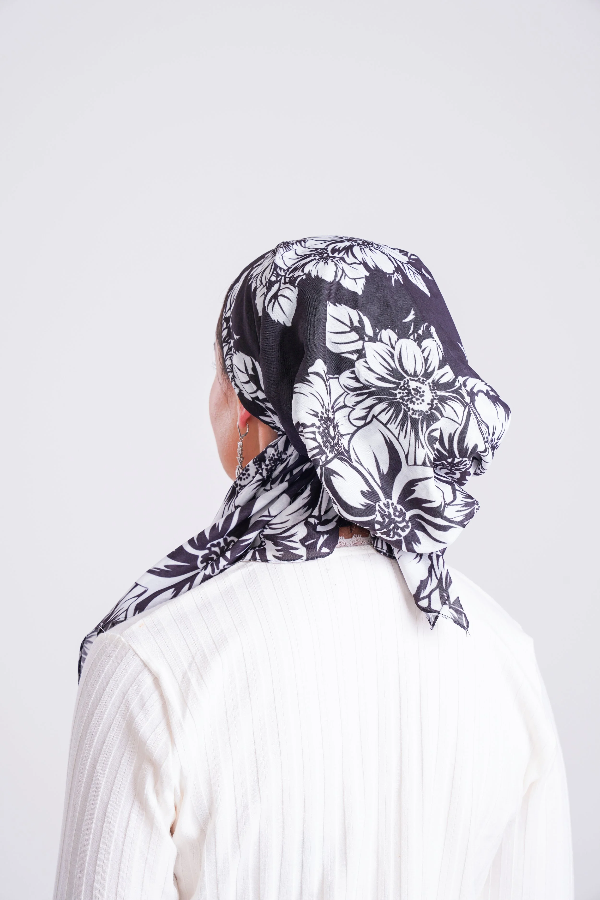 A woman wearing a black and white floral headscarf and a white top, facing away from the camera against a plain white background.