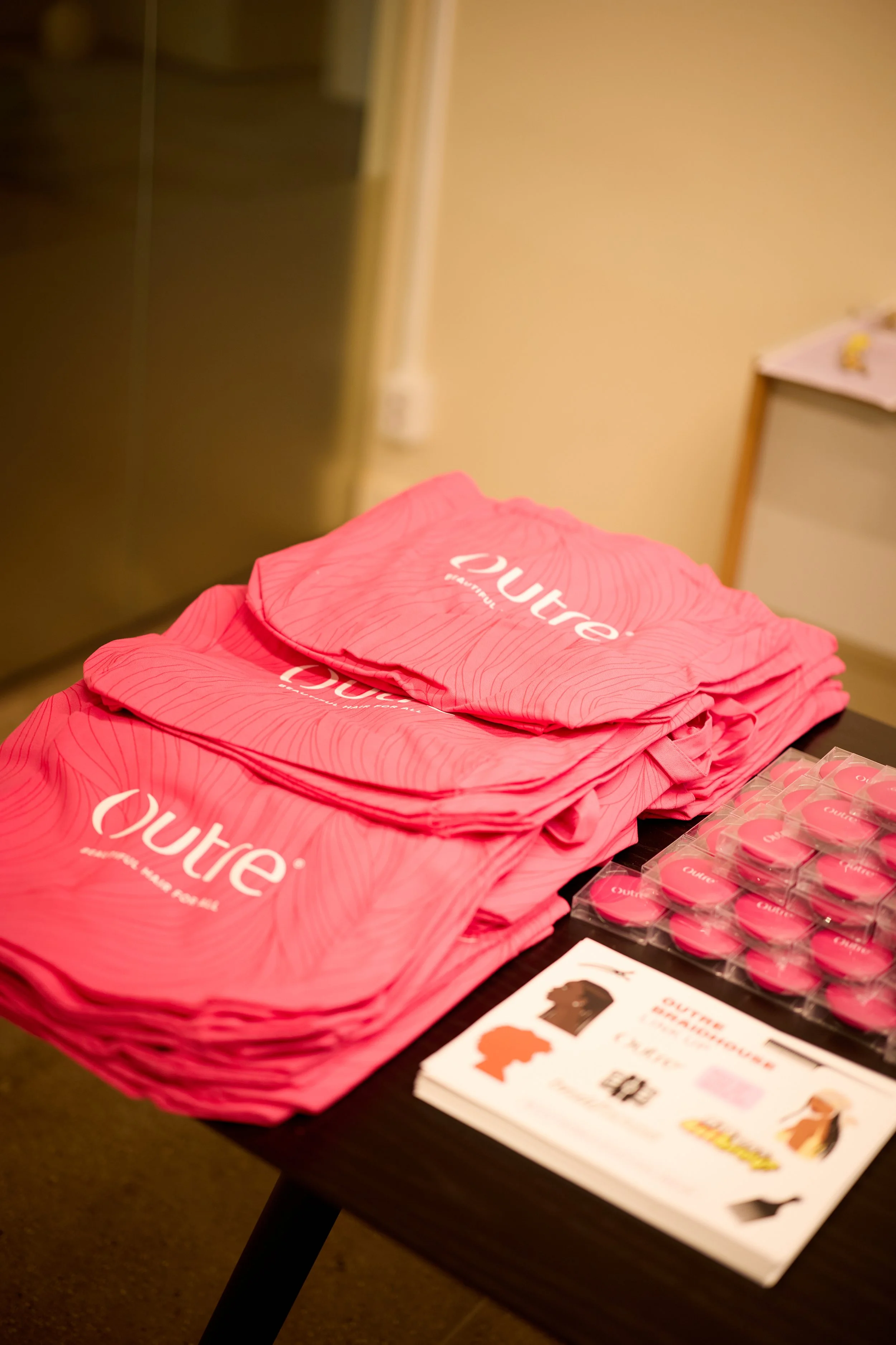 Pink drawstring bags with the white logo of Outre, a makeup brand, are stacked on a table. Next to the bags are pink buttons and promotional cards.