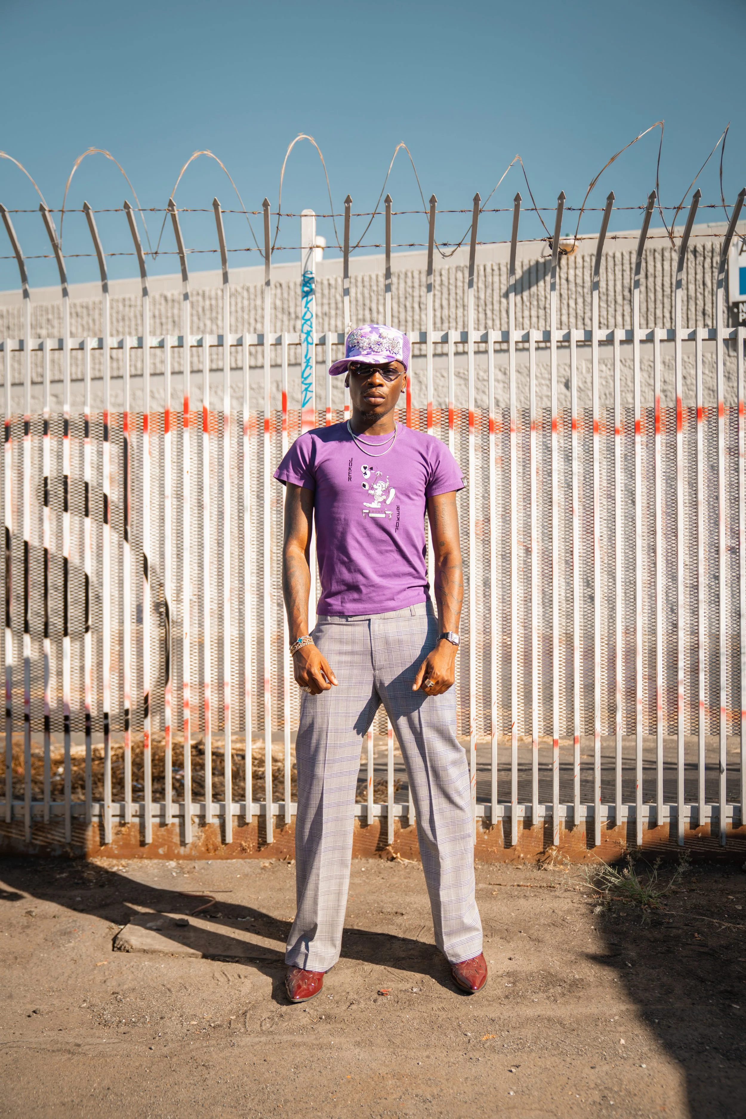 A person standing outdoors in front of a metal fence with barbed wire under a clear blue sky. They are wearing a purple T-shirt, patterned gray pants, a purple hat, sunglasses, and jewelry.