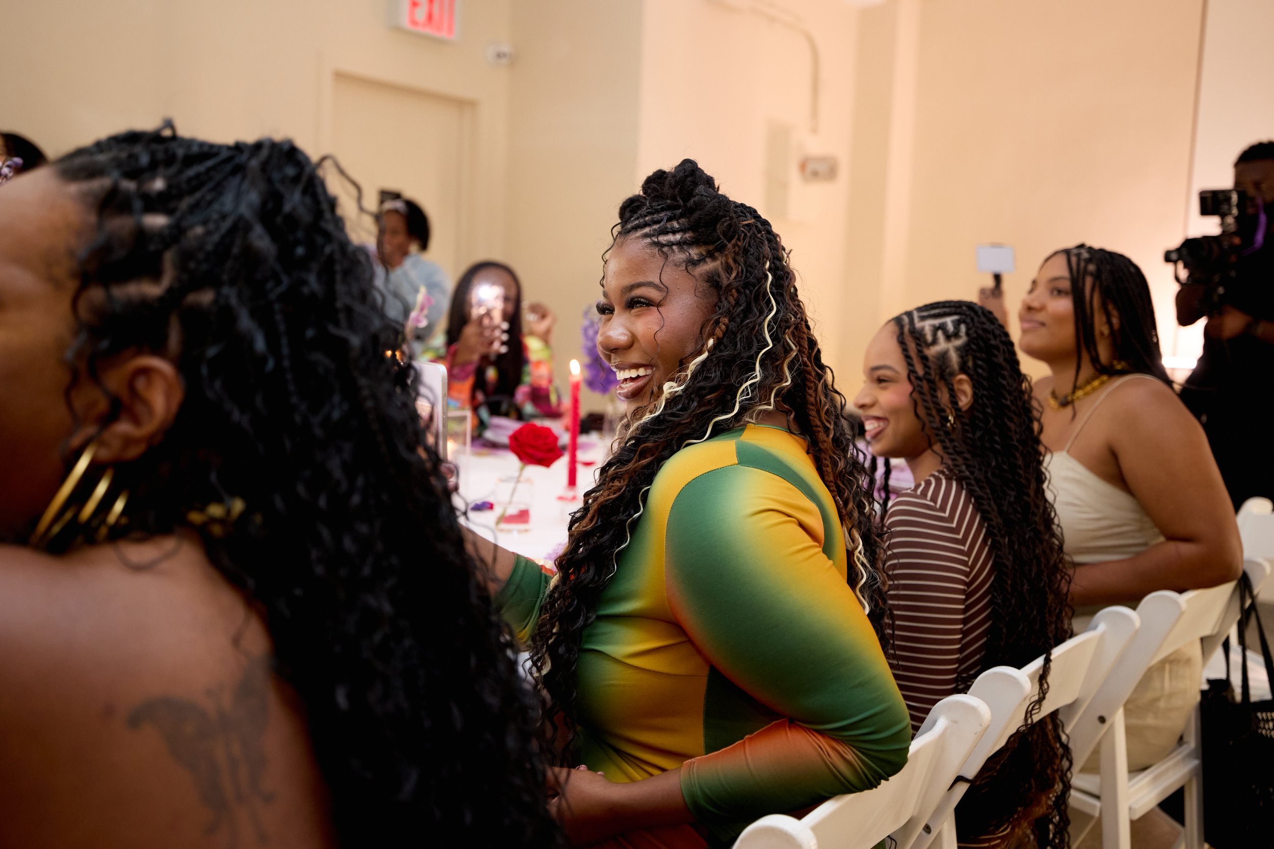 A group of women sitting at a table, smiling and enjoying a celebration, with one woman in the foreground wearing a colorful dress with green, yellow, and orange hues.