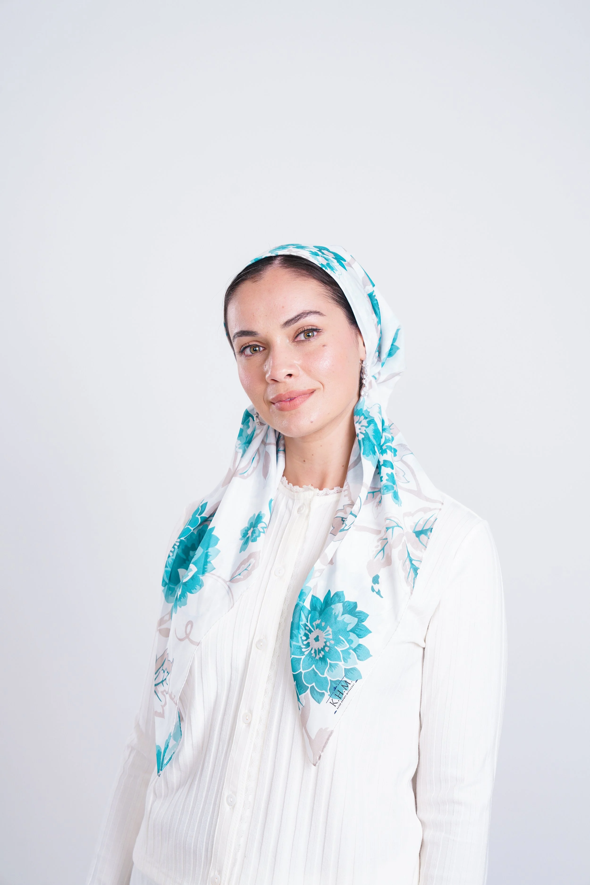 A woman with dark hair tied back, wearing a white blouse and a white headscarf with blue floral patterns, smiling at the camera against a plain white background.
