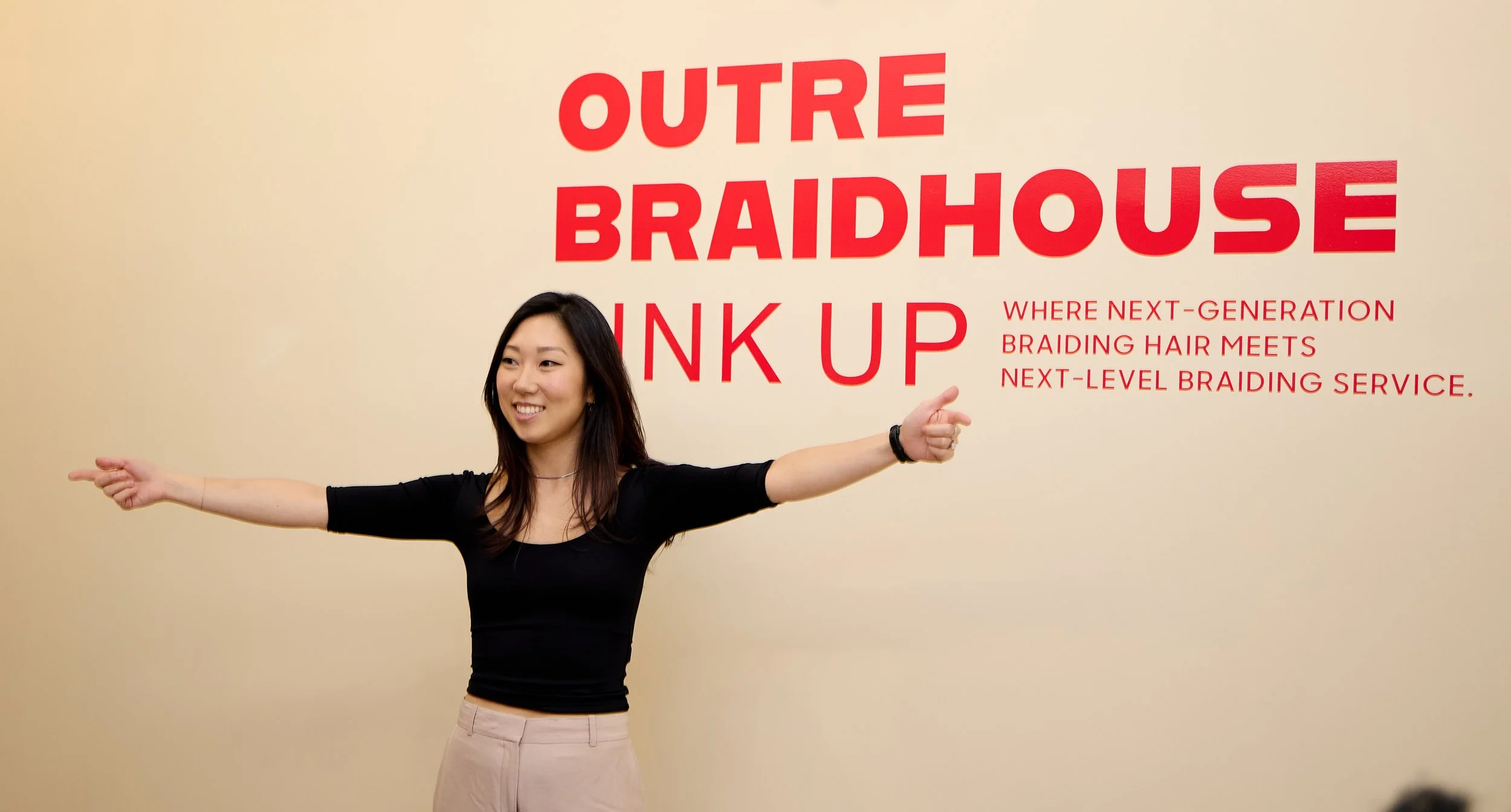A woman smiling with arms outstretched standing in front of a wall with red text advertising "Outre Braidhouse Ink Up," a next-generation braiding hair service.
