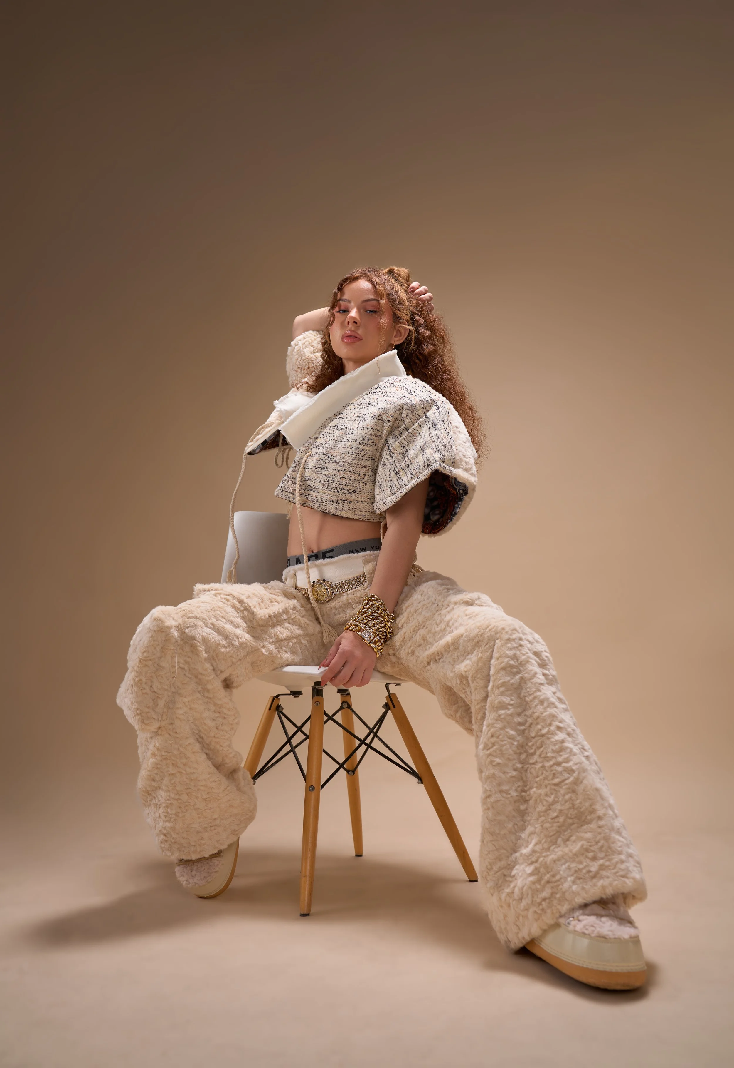A woman with curly hair seated on a modern wooden chair with metal supports, dressed in a beige layered outfit with baggy fleece pants and a cropped top, posing confidently in front of a beige background.