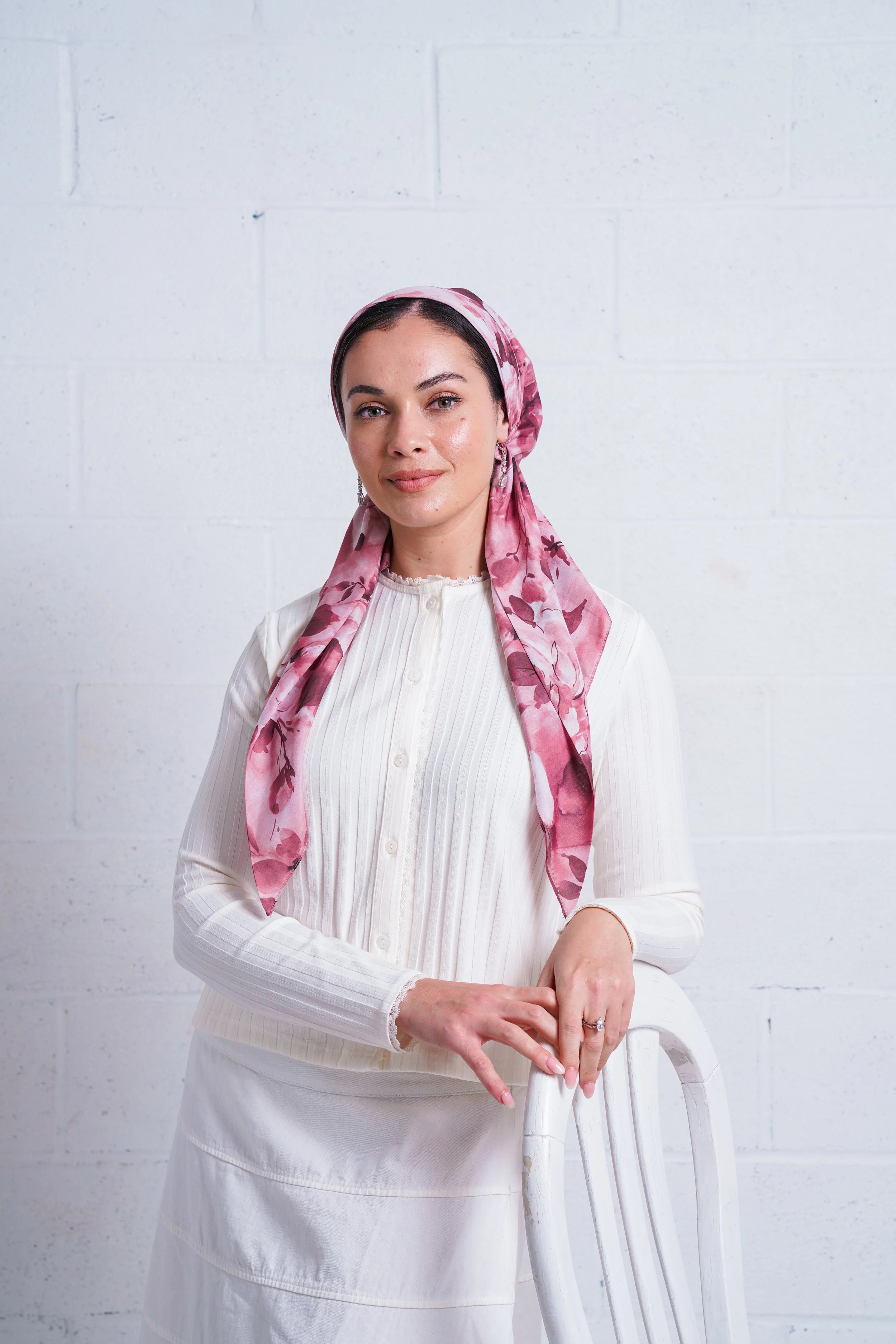 A woman with dark hair wearing a pink patterned headscarf and a white button-up sweater standing against a white brick wall, leaning on a white chair.