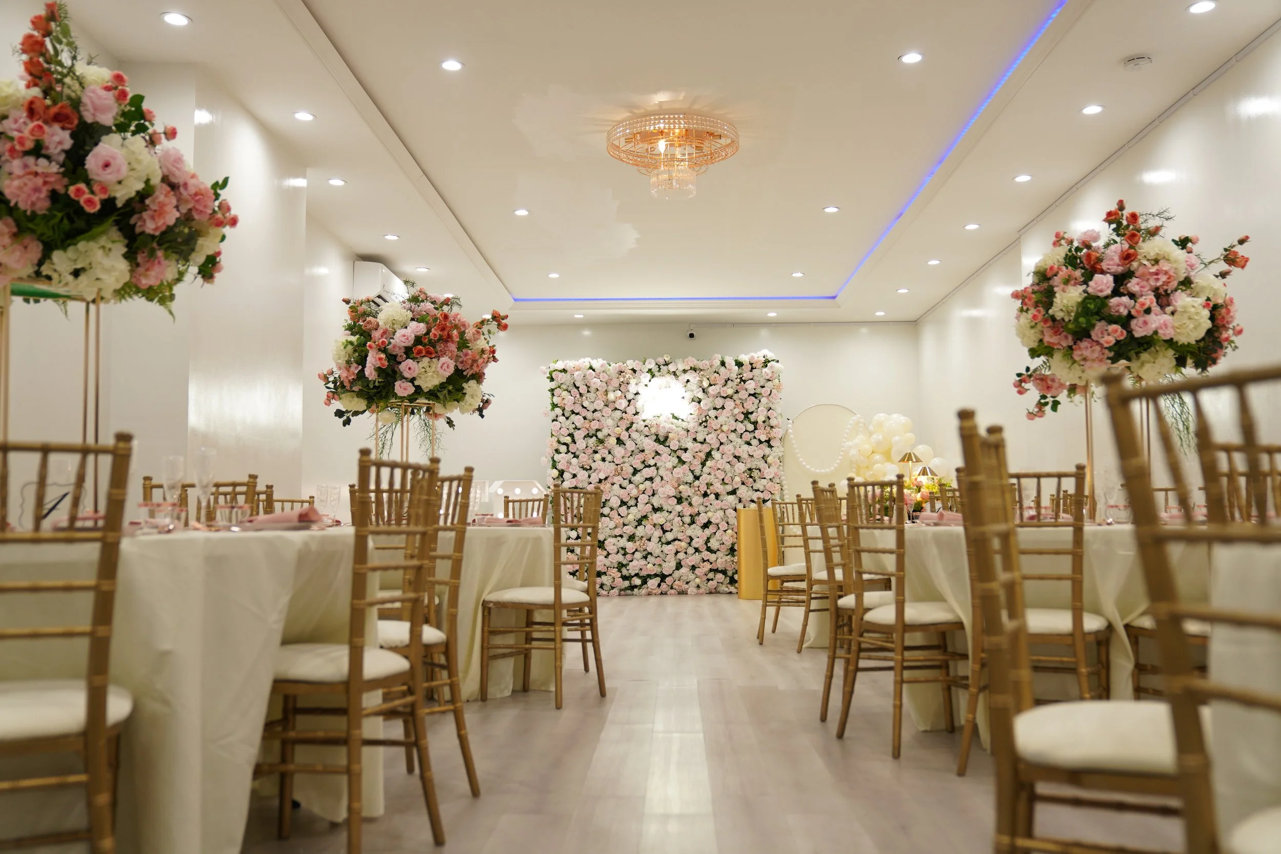 Elegant banquet hall decorated for a wedding reception with round tables, gold Chiavari chairs, floral centerpieces, and a large flower wall at the back.