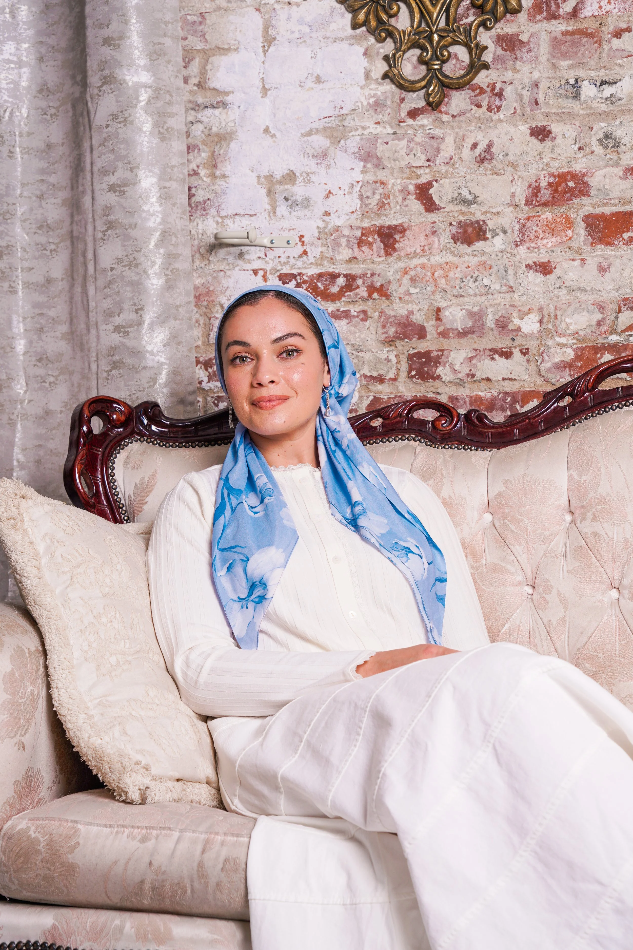A woman sitting on an ornate vintage cream-colored sofa with floral upholstery, against a rustic brick wall backdrop. She is wearing a white outfit, a blue patterned headscarf, and has a calming expression.