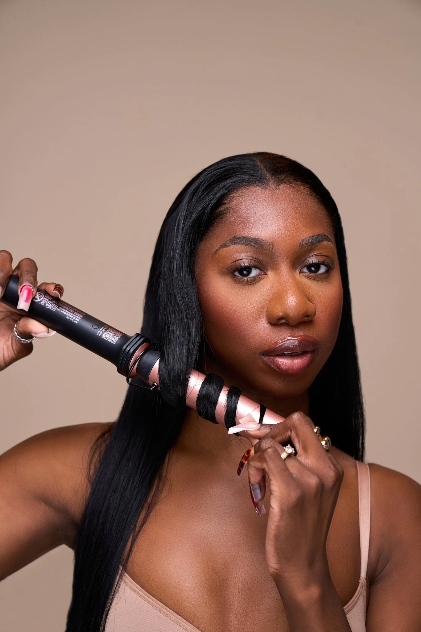 A woman with long black hair is holding a curling iron wrapped with hair and preparing to curl her hair. She is wearing a beige top and has rings on her fingers.