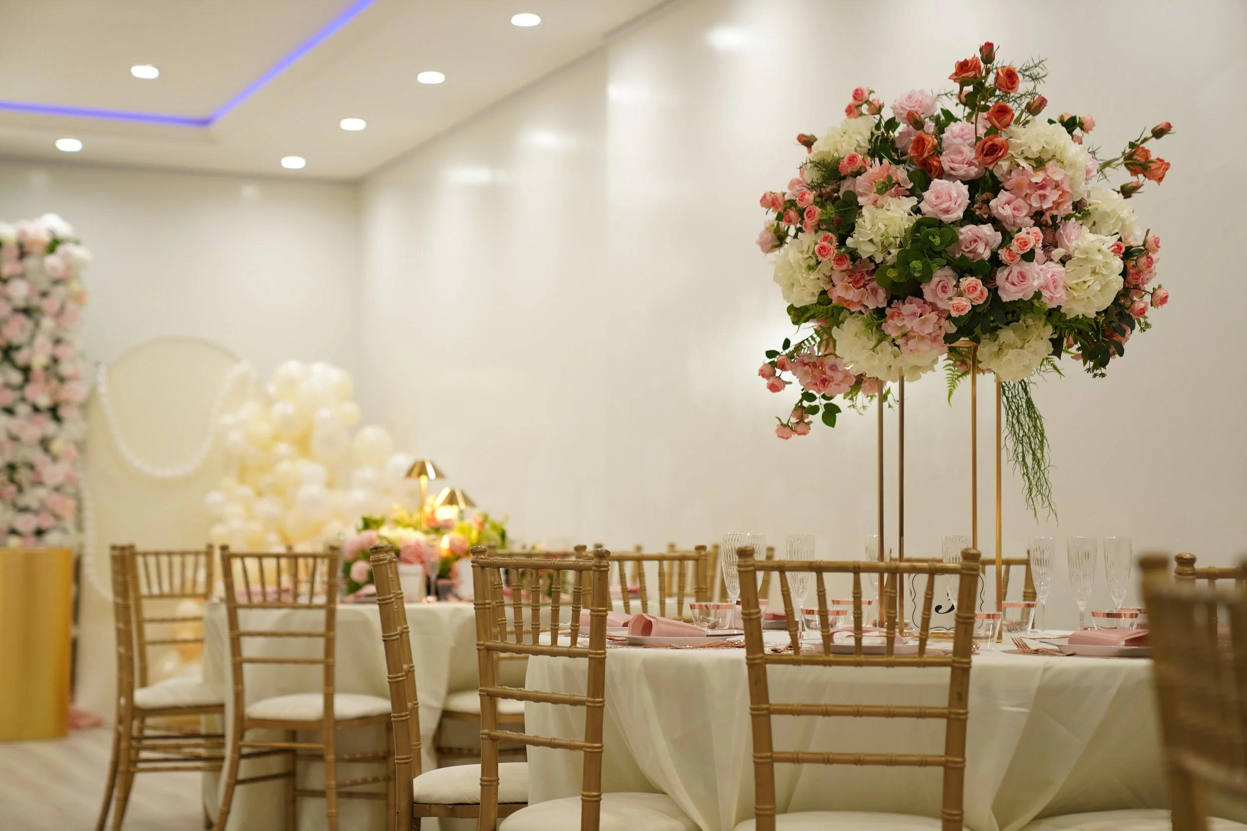 Elegant banquet table with a large floral centerpiece, set with glasses, pink napkins, and gold chairs in a decorated event space.