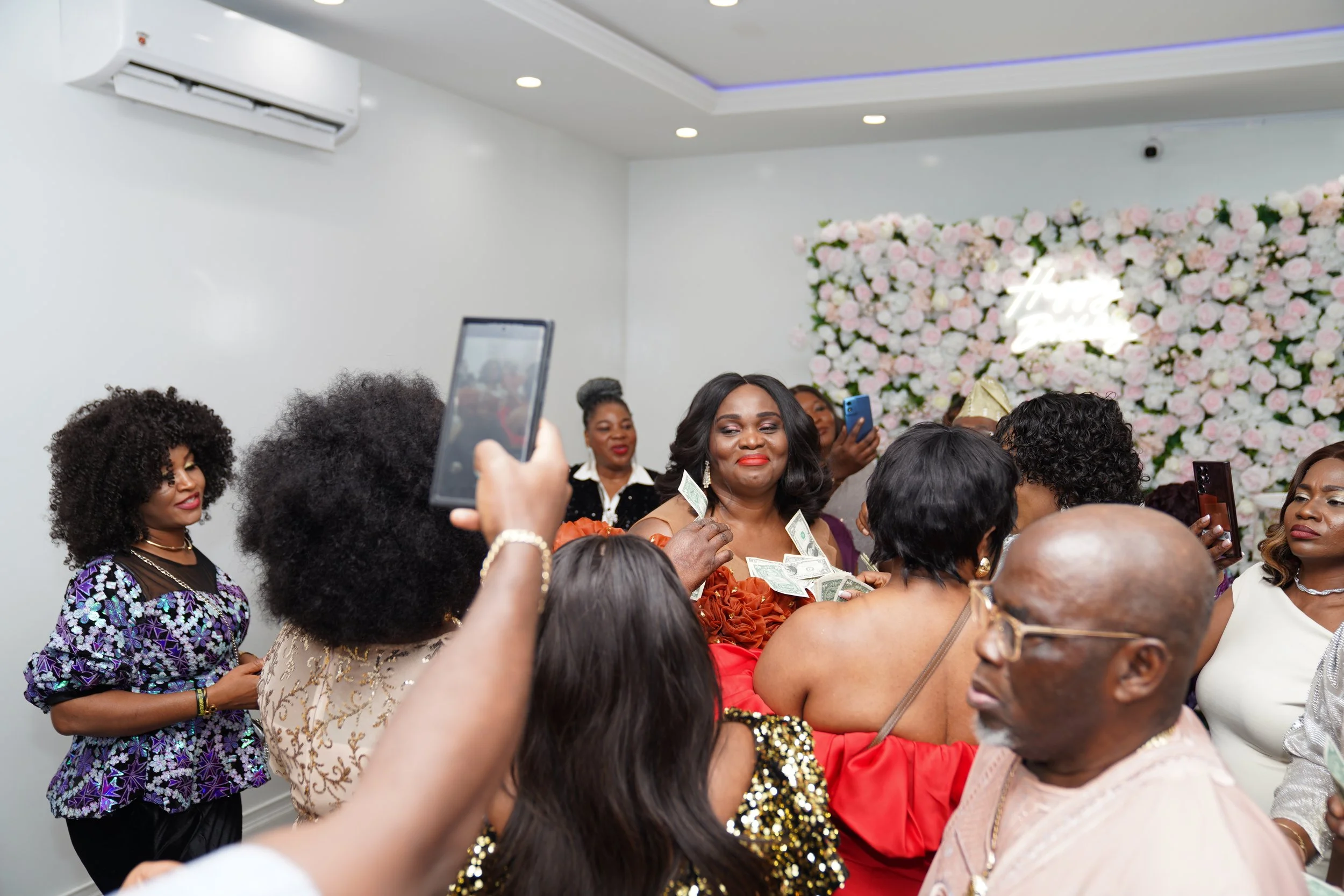 A group of people at a celebration, with a woman in an orange dress throwing money into the air, surrounded by others dressed in formal attire, with a floral backdrop and neon sign in the background.