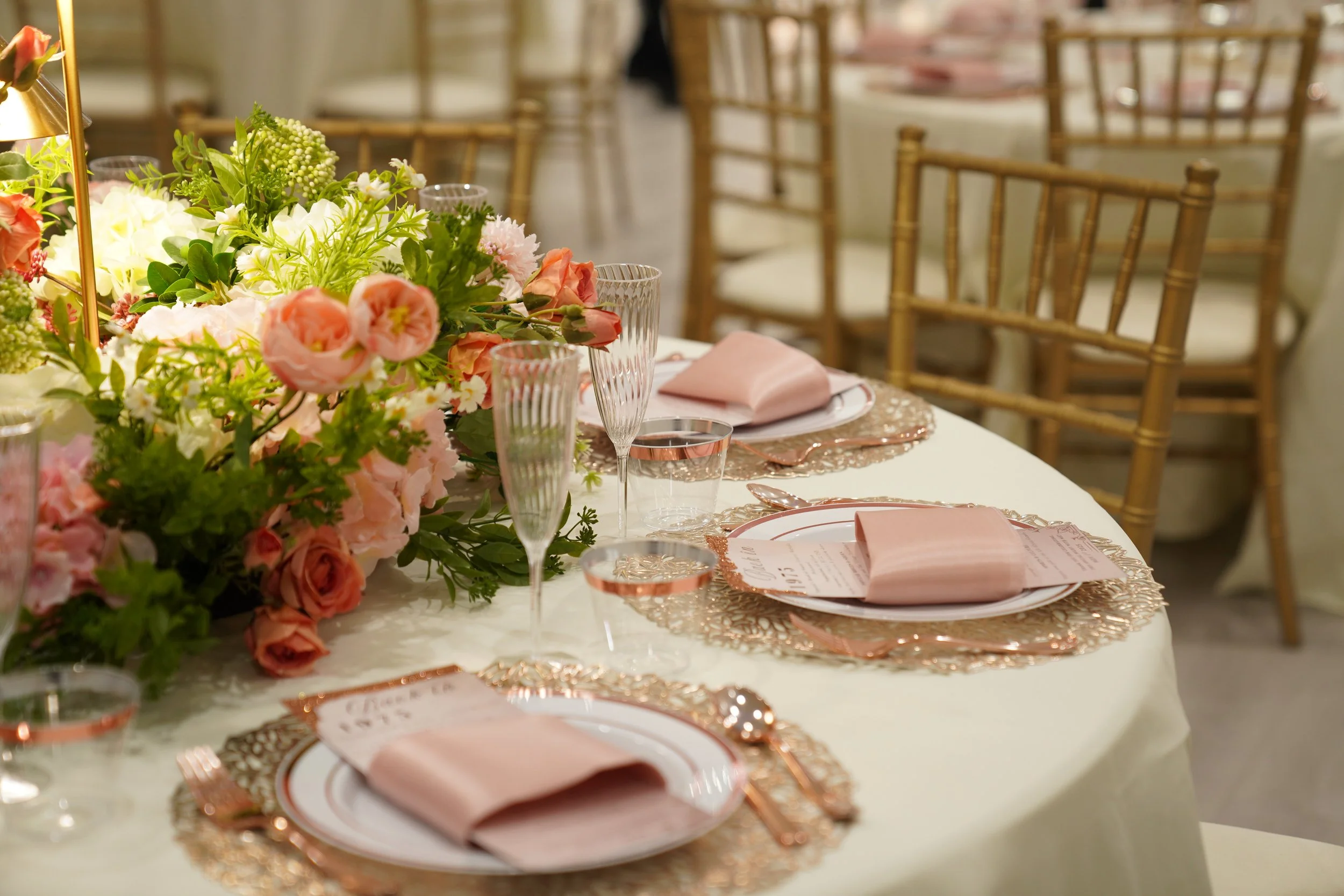 Elegant banquet table decorated with a large pink and white floral centerpiece, pink napkins on white plates, gold-colored cutlery, and glassware, set for a formal event or wedding reception.