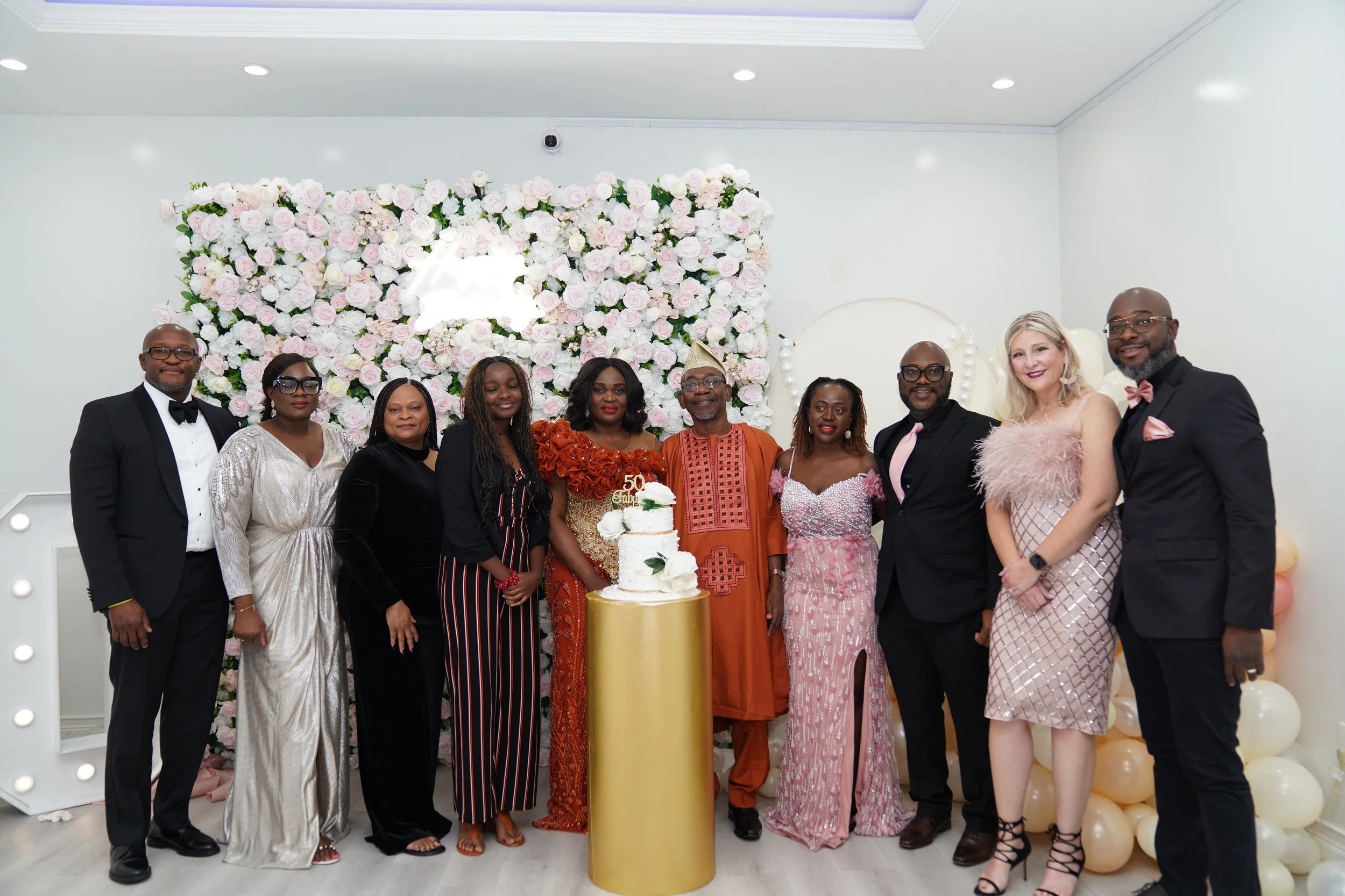 A group of twelve people dressed formally, standing together in front of a backdrop of pink and white roses, celebrating a 50th anniversary with a cake in the center. The setting appears to be indoors, with white walls and ceiling lights.