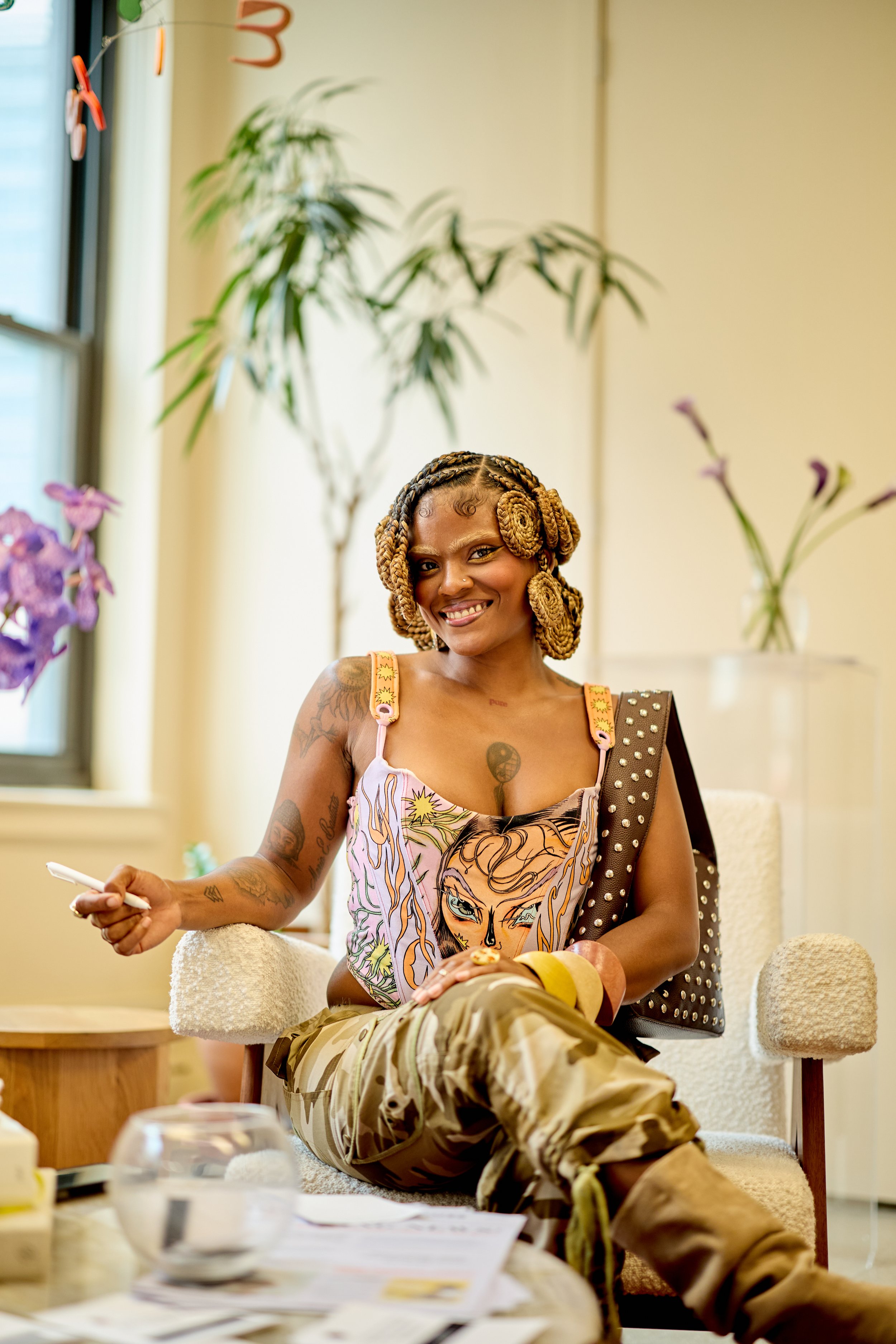 A smiling woman with braided hair, tattoos, and a colorful tank top featuring a woman's face, sitting comfortably on a beige chair in a well-lit room with plants and purple flowers.