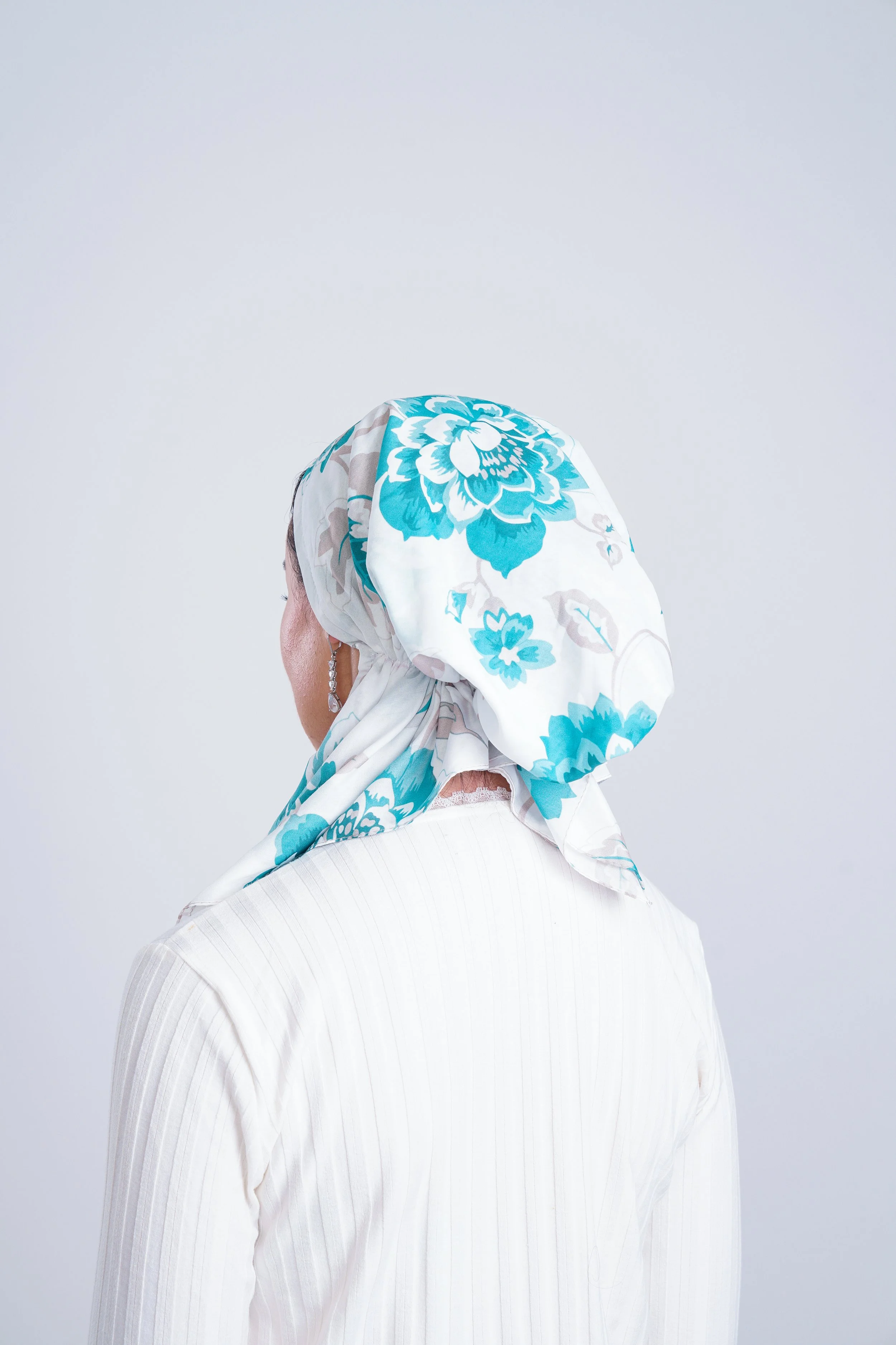 Woman wearing a white shirt and a floral headscarf with blue and turquoise flowers, facing away from the camera.