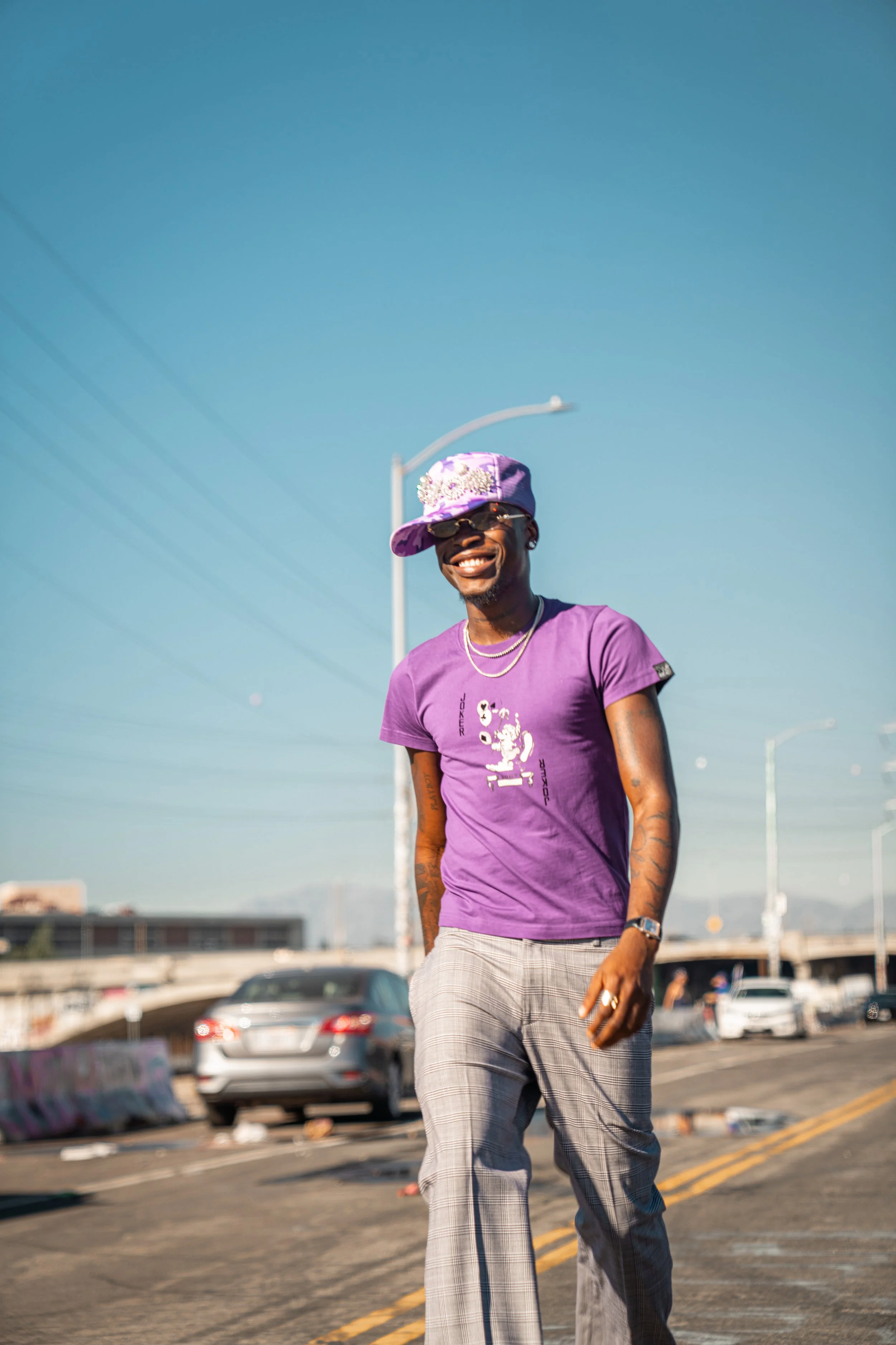 A man walking on a city street during the daytime, wearing a purple T-shirt, plaid trousers, a purple cap, sunglasses, and jewelry, smiling.