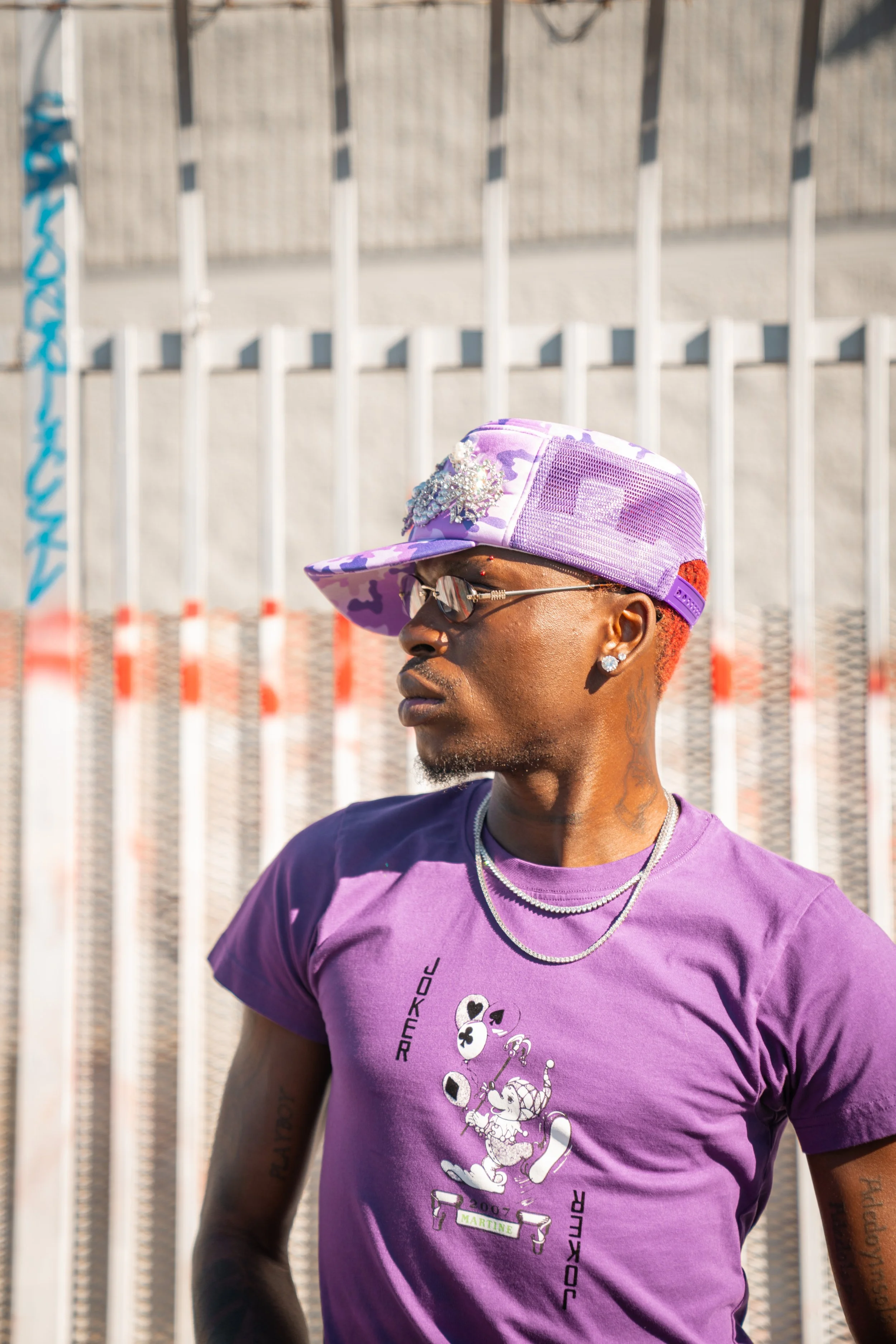 A young man wearing a purple graphic t-shirt, purple camouflage cap with decorative embellishment, sunglasses, earrings, and layered chains, standing in front of a fence with vertical white bars and red-orange caution tape.