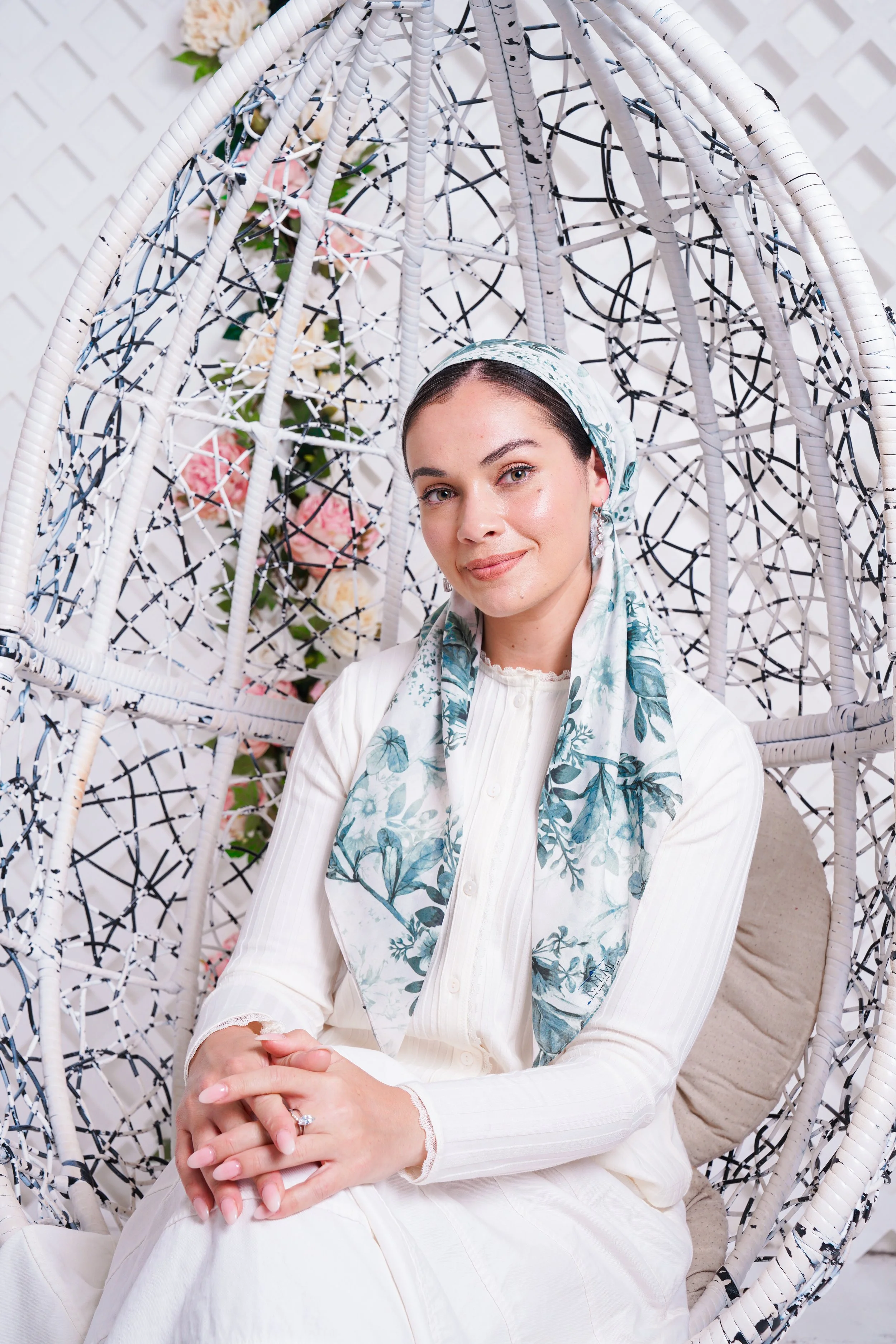 A woman sitting on an ornate white woven chair with a lattice pattern. She is wearing a light-colored outfit with a floral scarf and has a ring on her finger. There are pink and white flowers in the background.