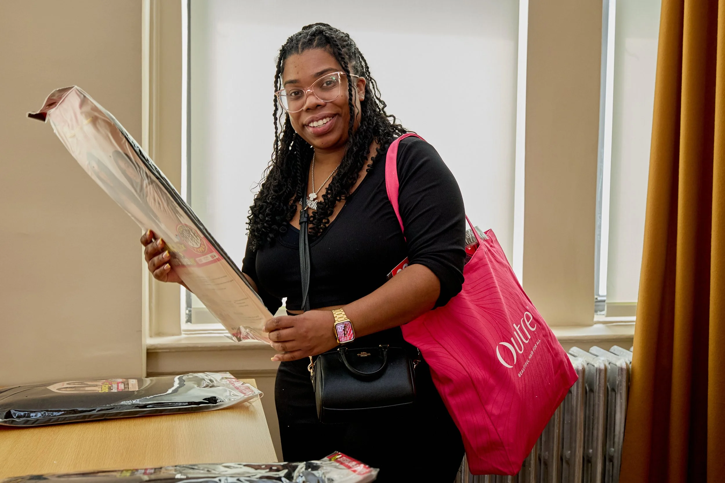 Woman with glasses, black top, pink tote bag, and smartwatch looking at packaged items on a table.