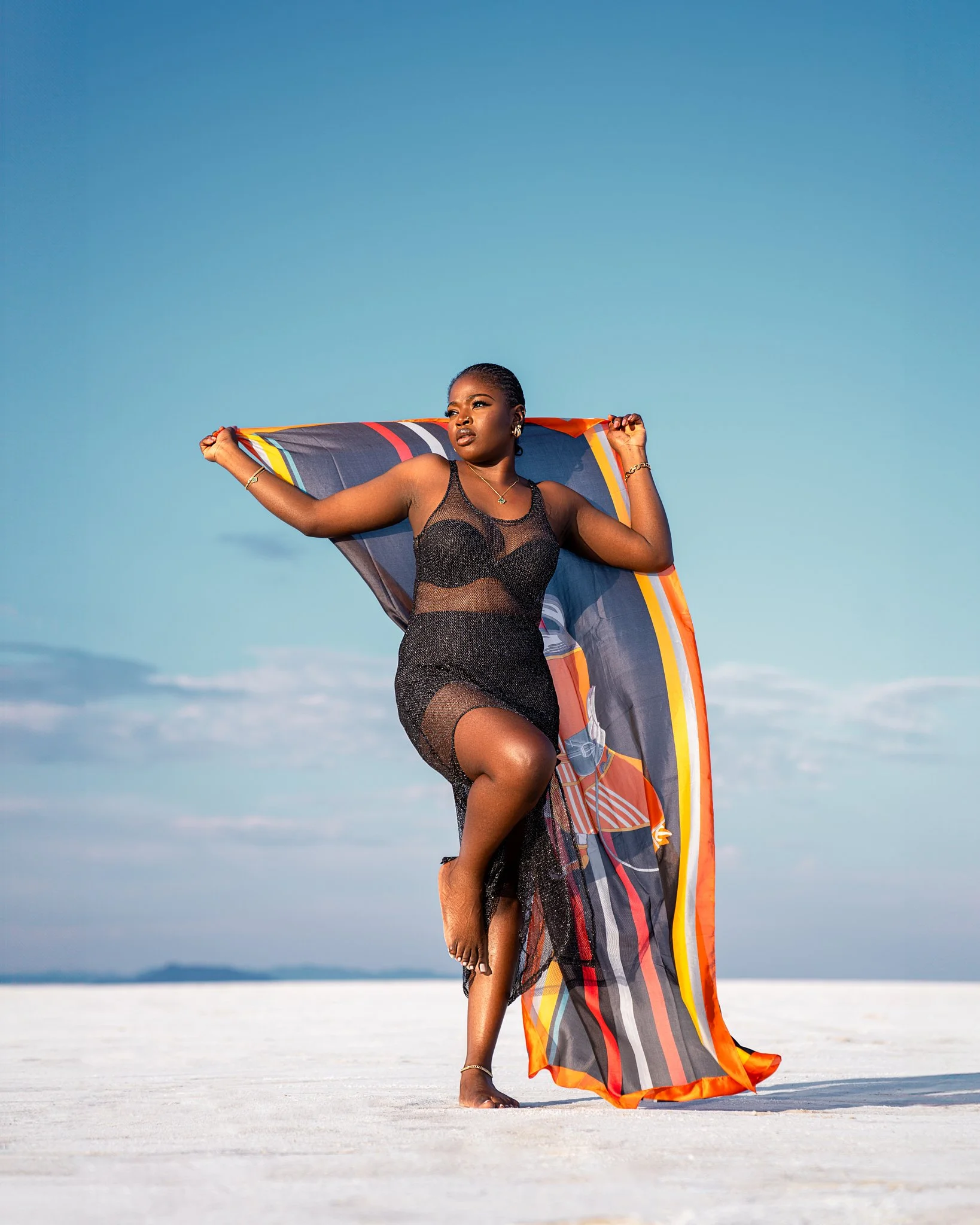 A woman in a black sheer dress stands on a white sandy beach, holding a colorful fabric behind her head against a clear blue sky.