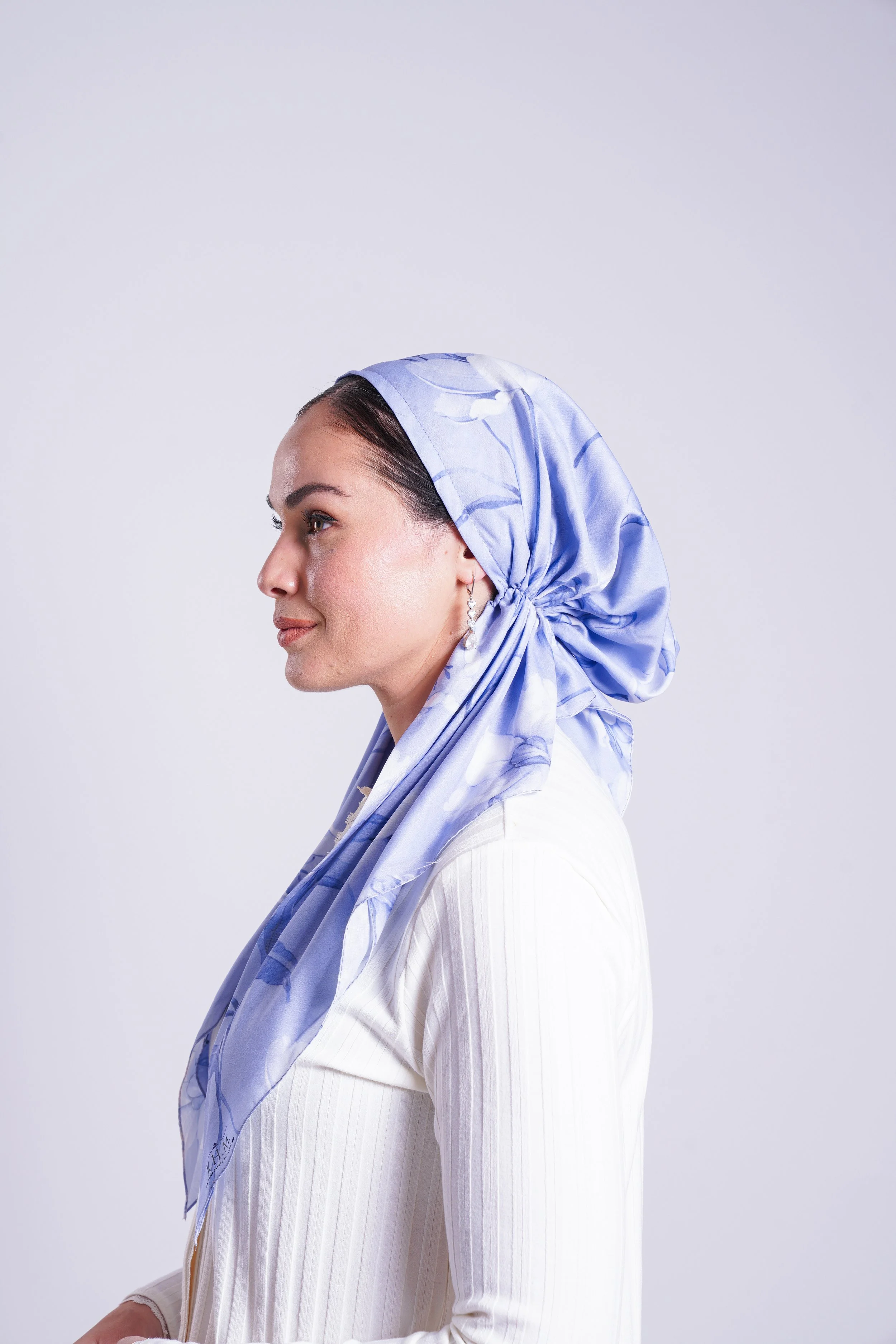 Profile of a woman with dark hair, wearing a light blue patterned headscarf and white top, against a plain background.