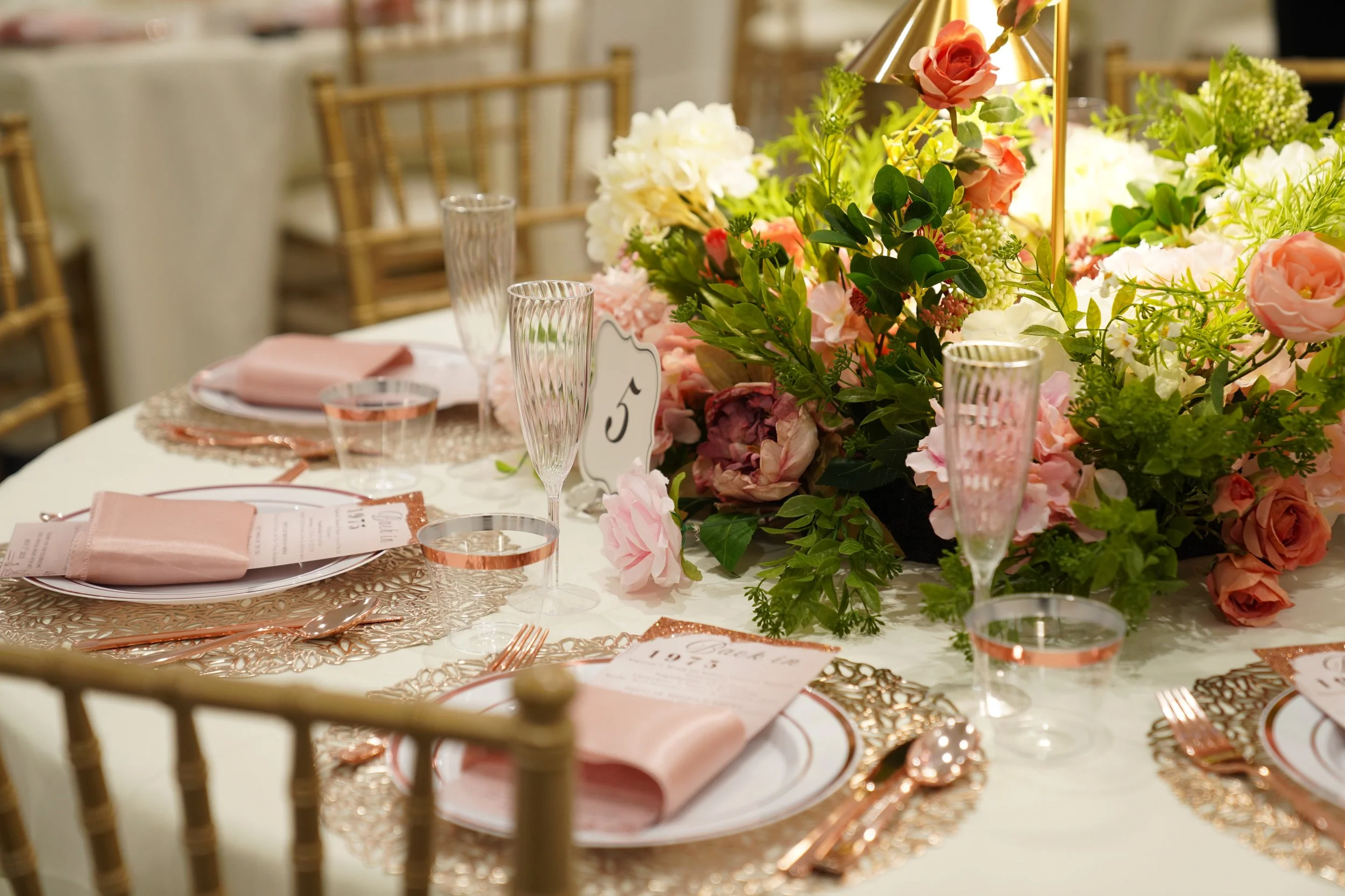 Elegant table setting with pink napkins, gold cutlery, floral centerpiece with pink and white flowers, and gold chairs in the background.