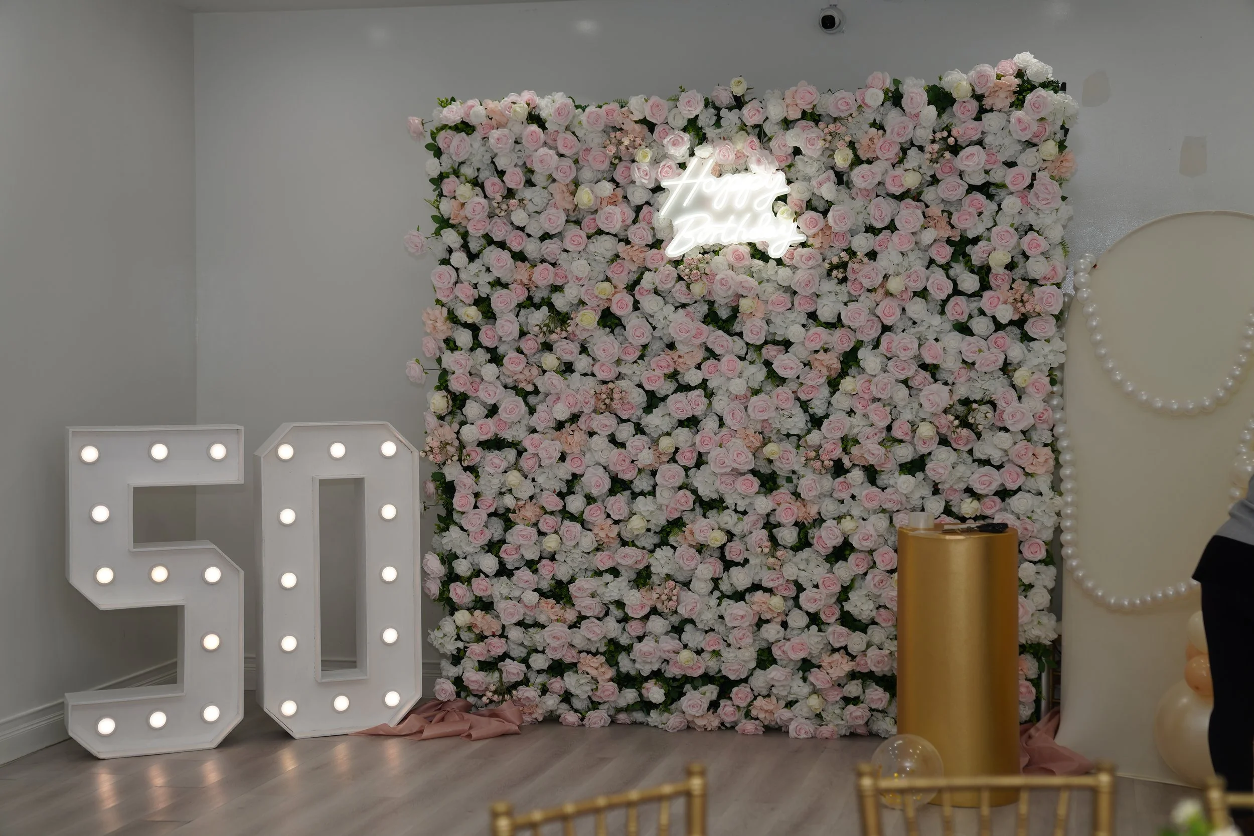 Decorative floral backdrop with pink and white roses, illuminated large number 50 on the left, and a neon sign that reads 'Happy Birthday' on the wall.