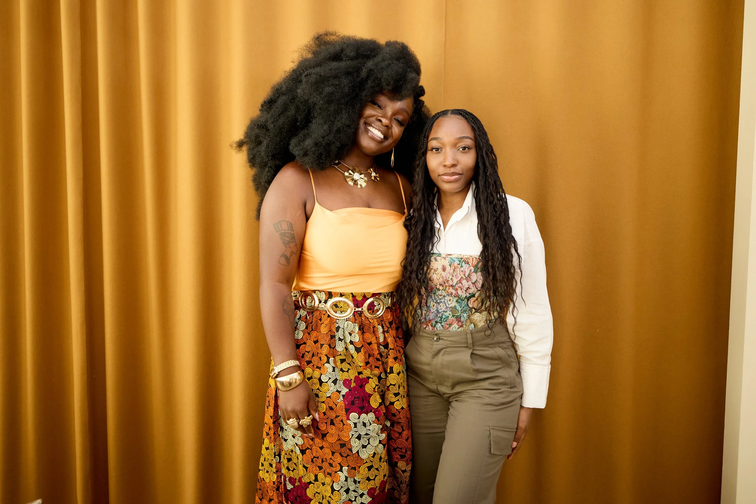 Two women stand together in front of a gold curtain, smiling at the camera. The woman on the left has dark curly hair and wears a yellow sleeveless top and a colorful patterned skirt. The woman on the right has long, dark, wavy hair, and is dressed i