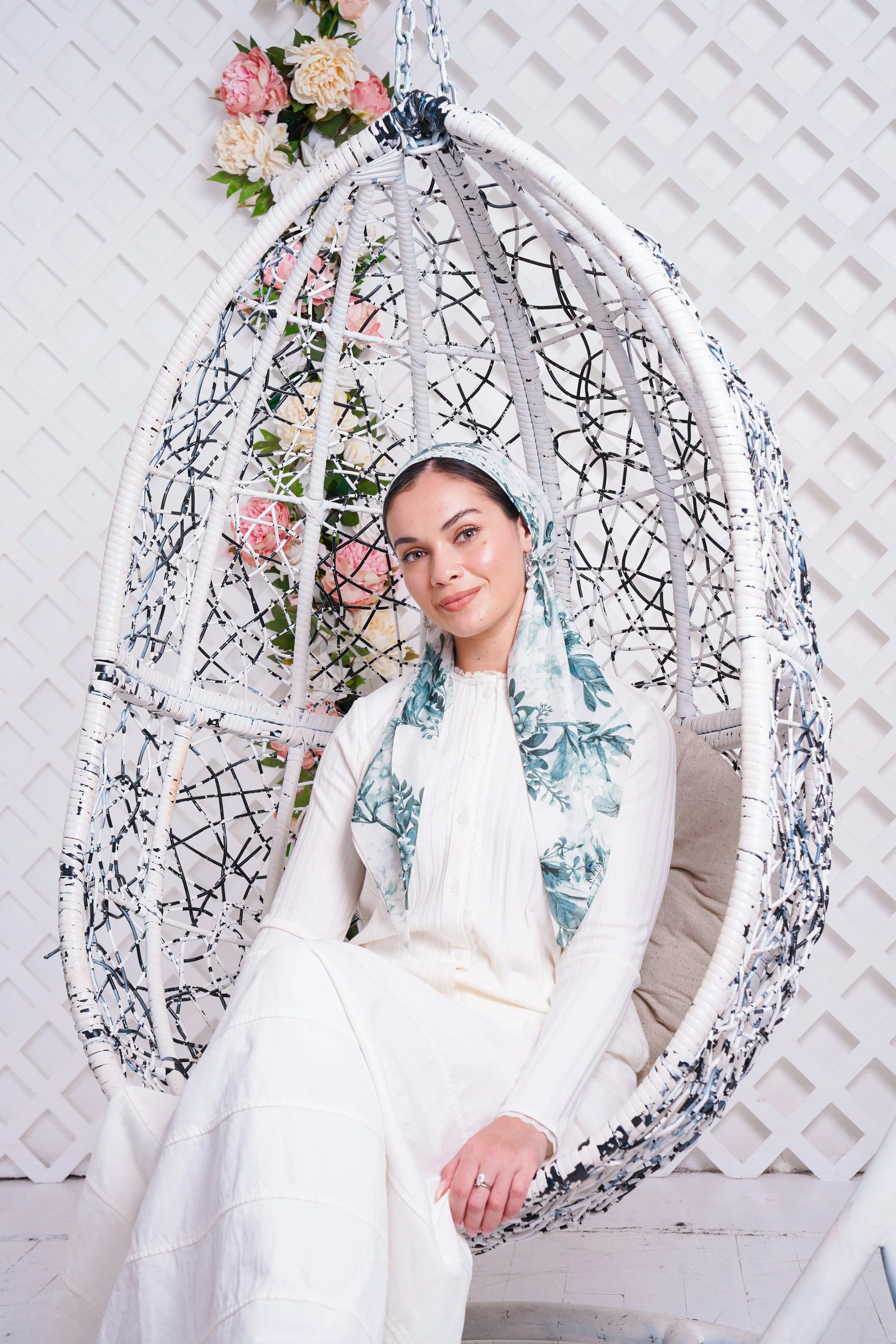 A woman sitting inside a hanging white wicker egg-shaped chair with a floral tapestry at the back, against a white lattice wall.