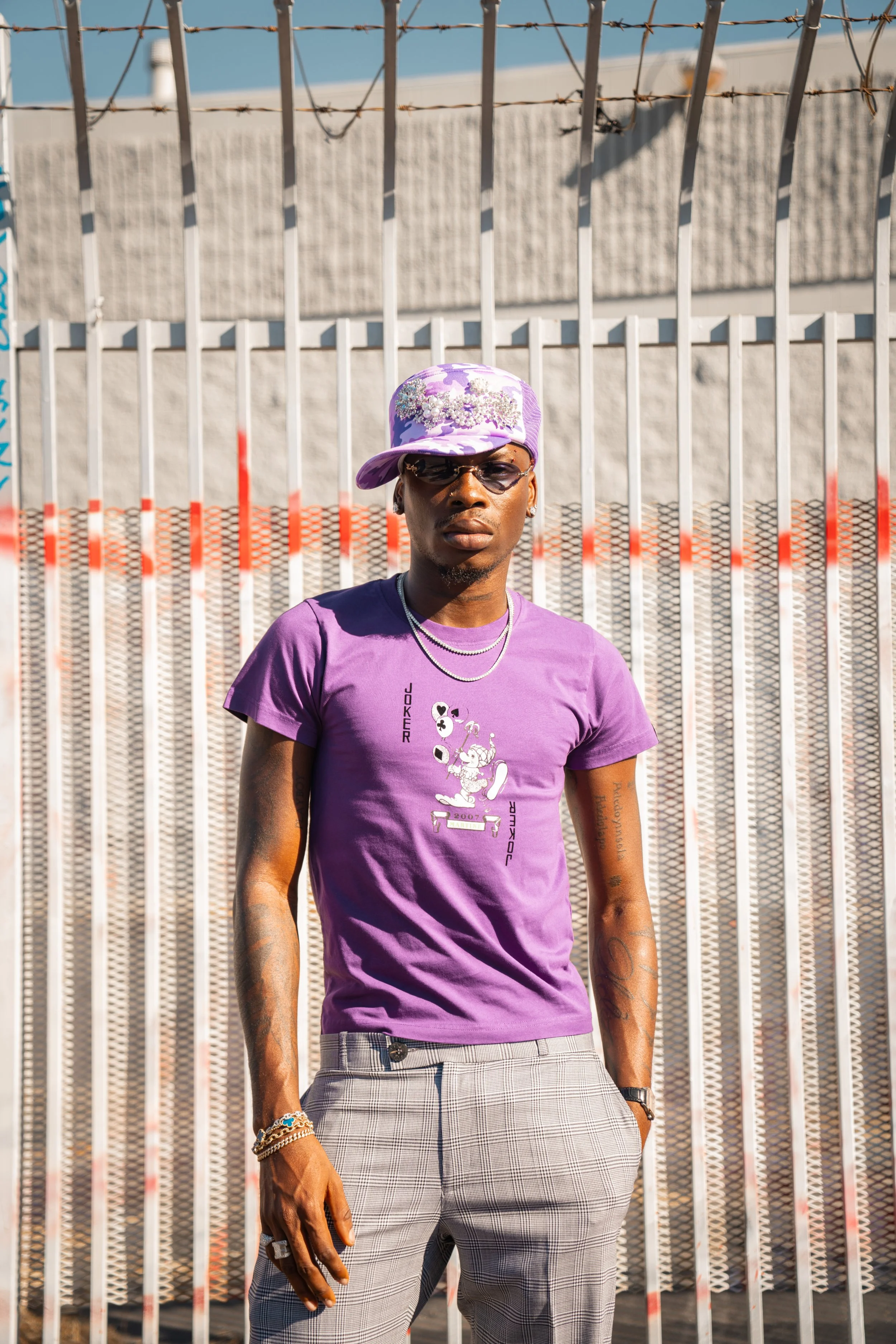 A stylish man in a purple t-shirt and plaid pants standing in front of a metal fence with barbed wire on top, wearing sunglasses, a purple cap, and jewelry.