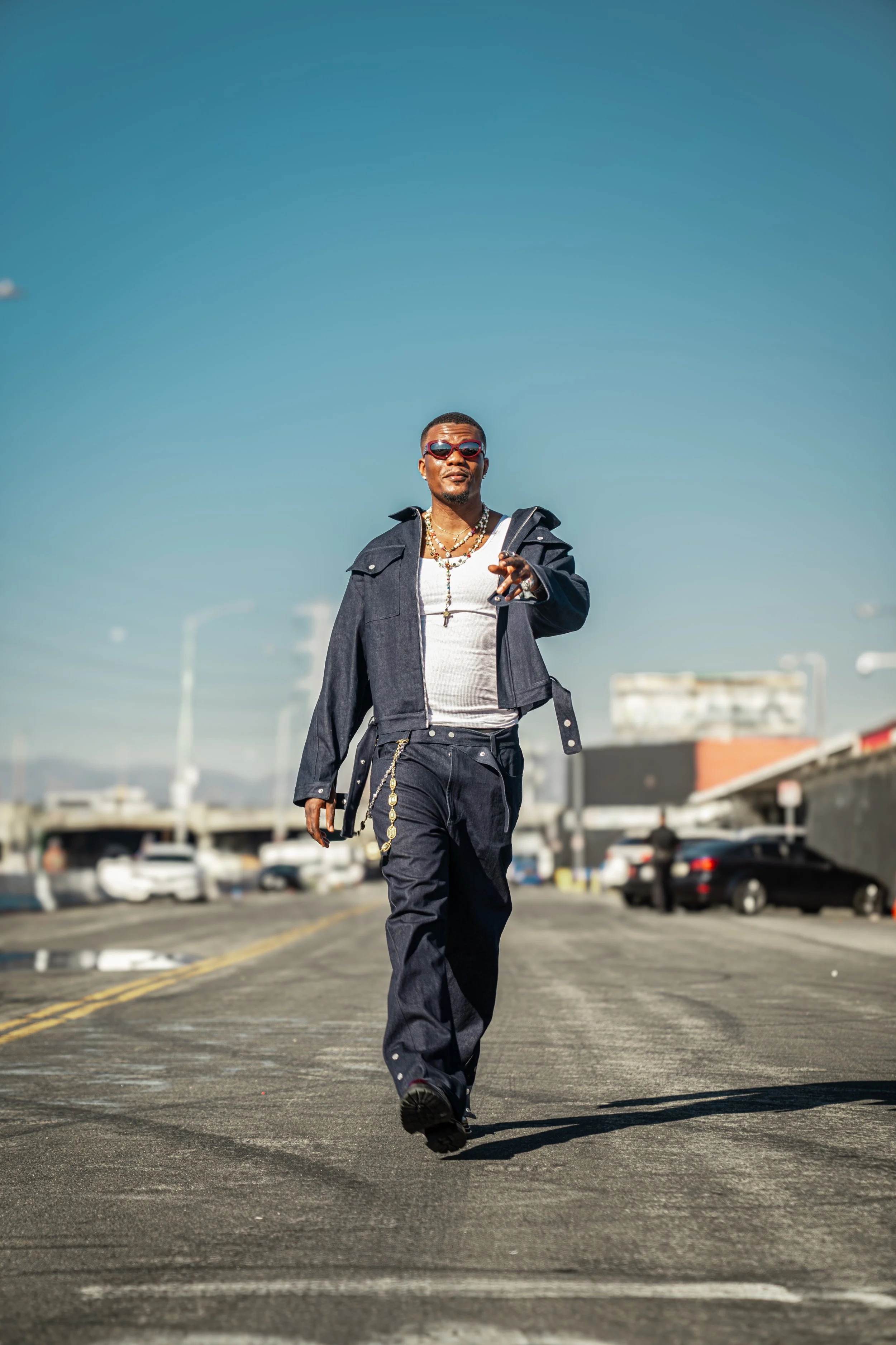 A man walking on a city street wearing sunglasses, a white tank top, a dark jacket, and loose dark pants with jewelry.