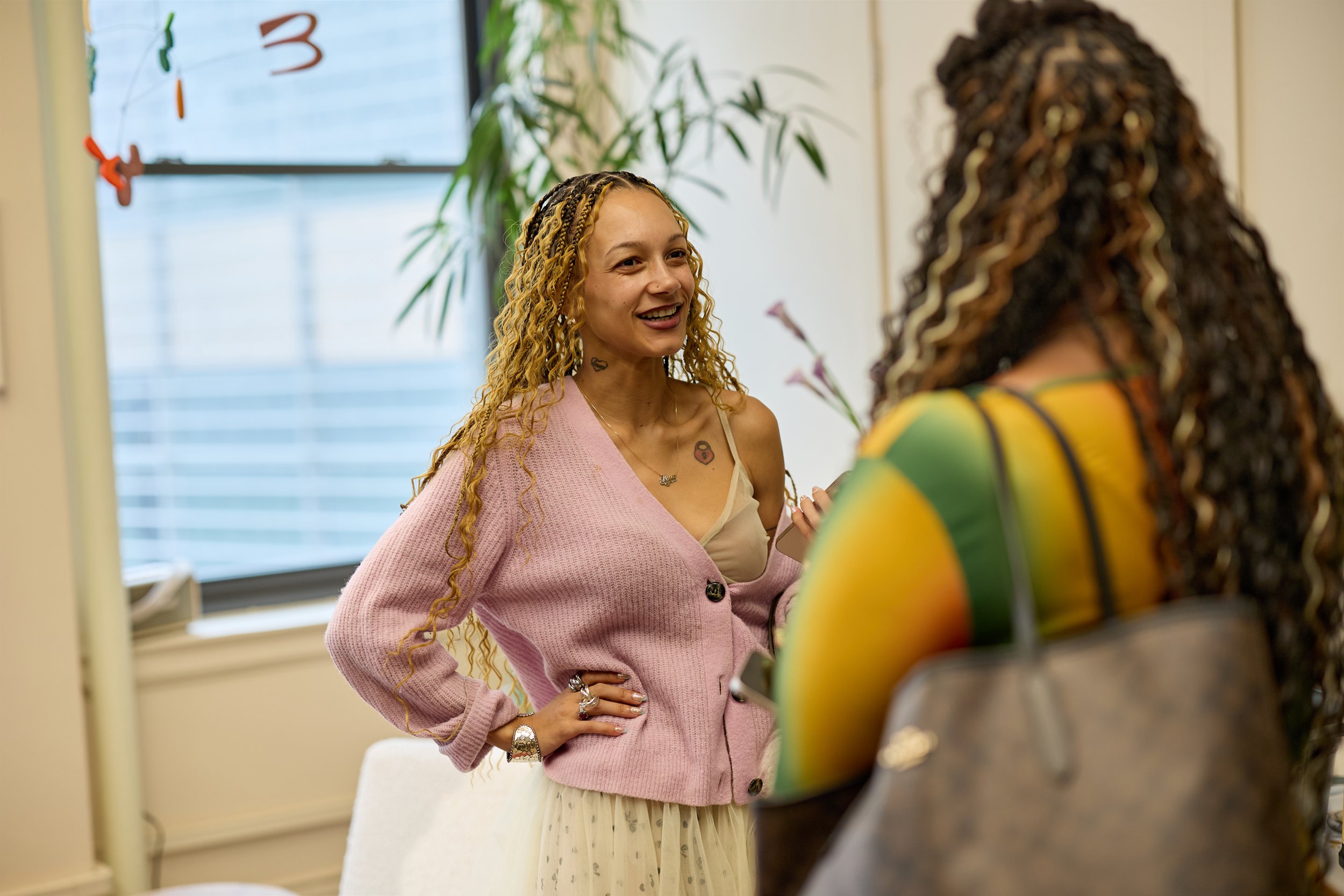 Two women having a conversation in an indoor setting. One woman with long, curly blonde hair is smiling and wearing a pink cardigan over a beige top, accessorized with rings and a watch. The other woman, whose back is to the camera, has long, curly d