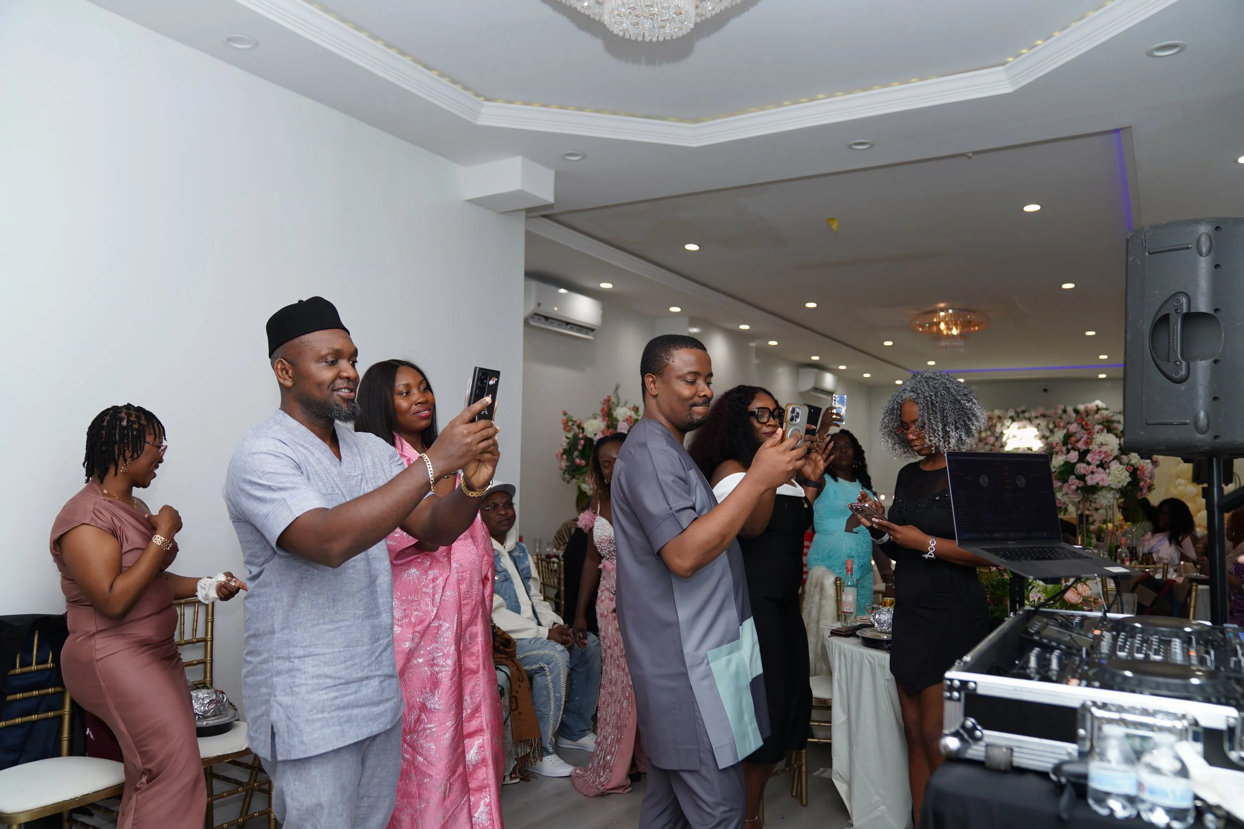 People at a celebration or event, some taking photos or videos with smartphones, in a decorated hall with floral arrangements and a DJ setup.