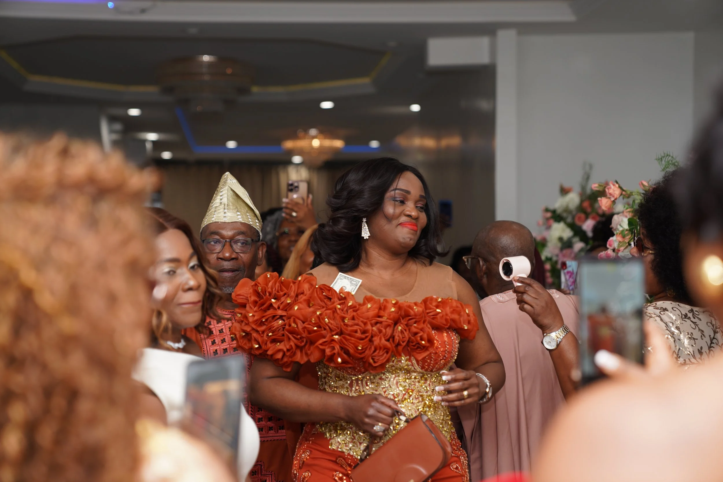 A group of people at a celebration, with a woman in the center wearing an elegant orange and gold dress with ruffled details and smiling slightly. Other attendees are dressed festively, some taking photos, in a decorated indoor setting.