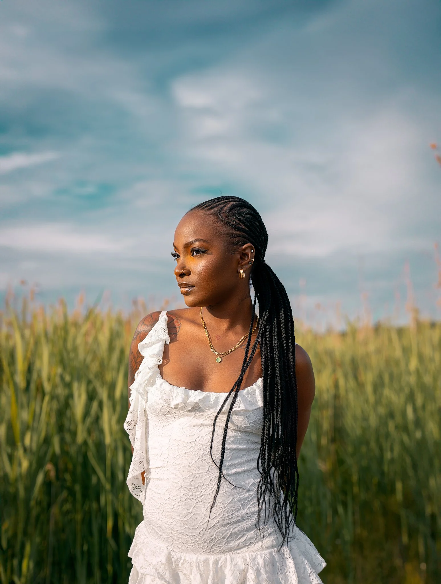 A woman with braided hair and earrings standing in a field of tall grass under a cloudy sky.