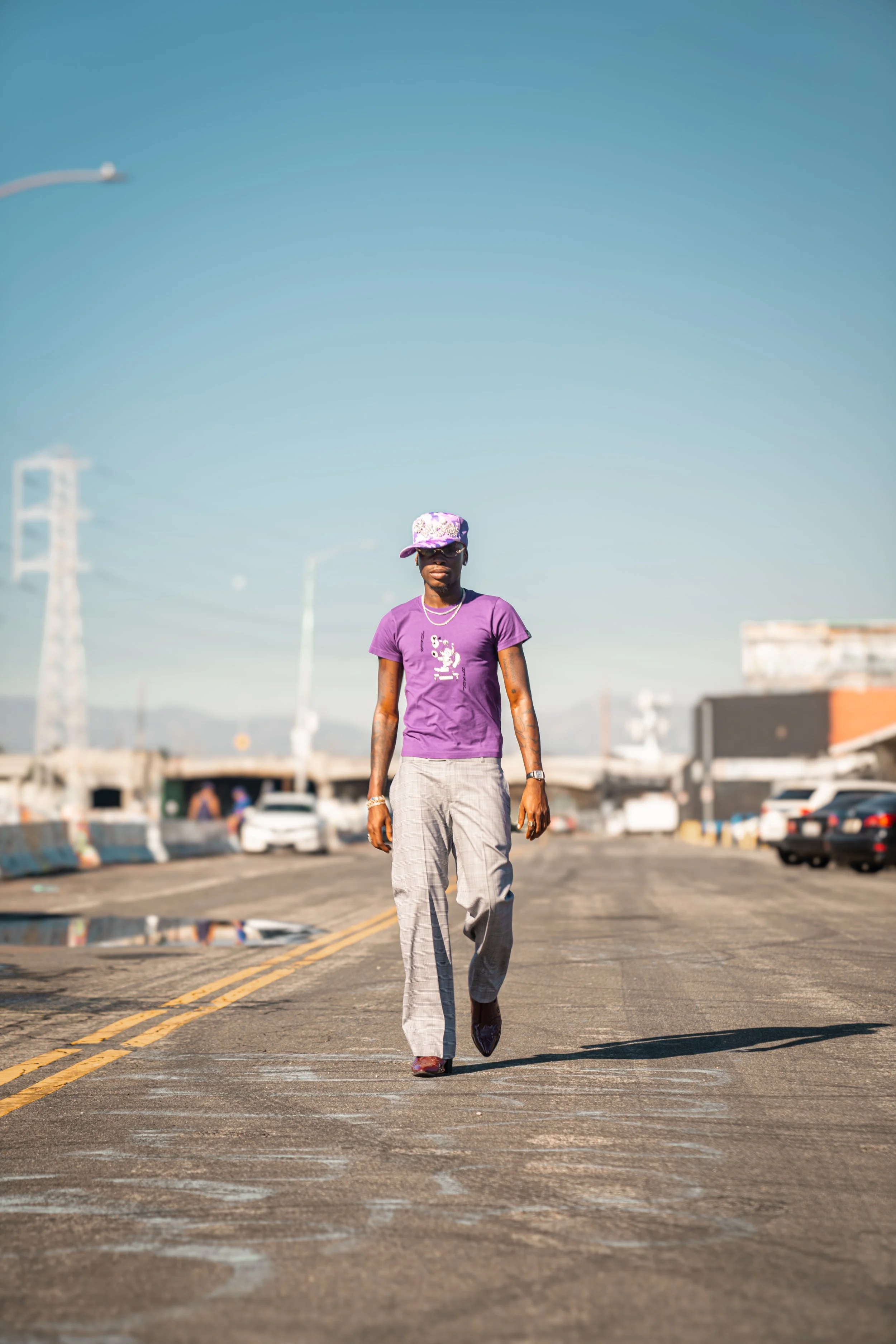 A woman walking on a paved road in an urban area with cars parked along the side and industrial buildings in the background under a clear blue sky.