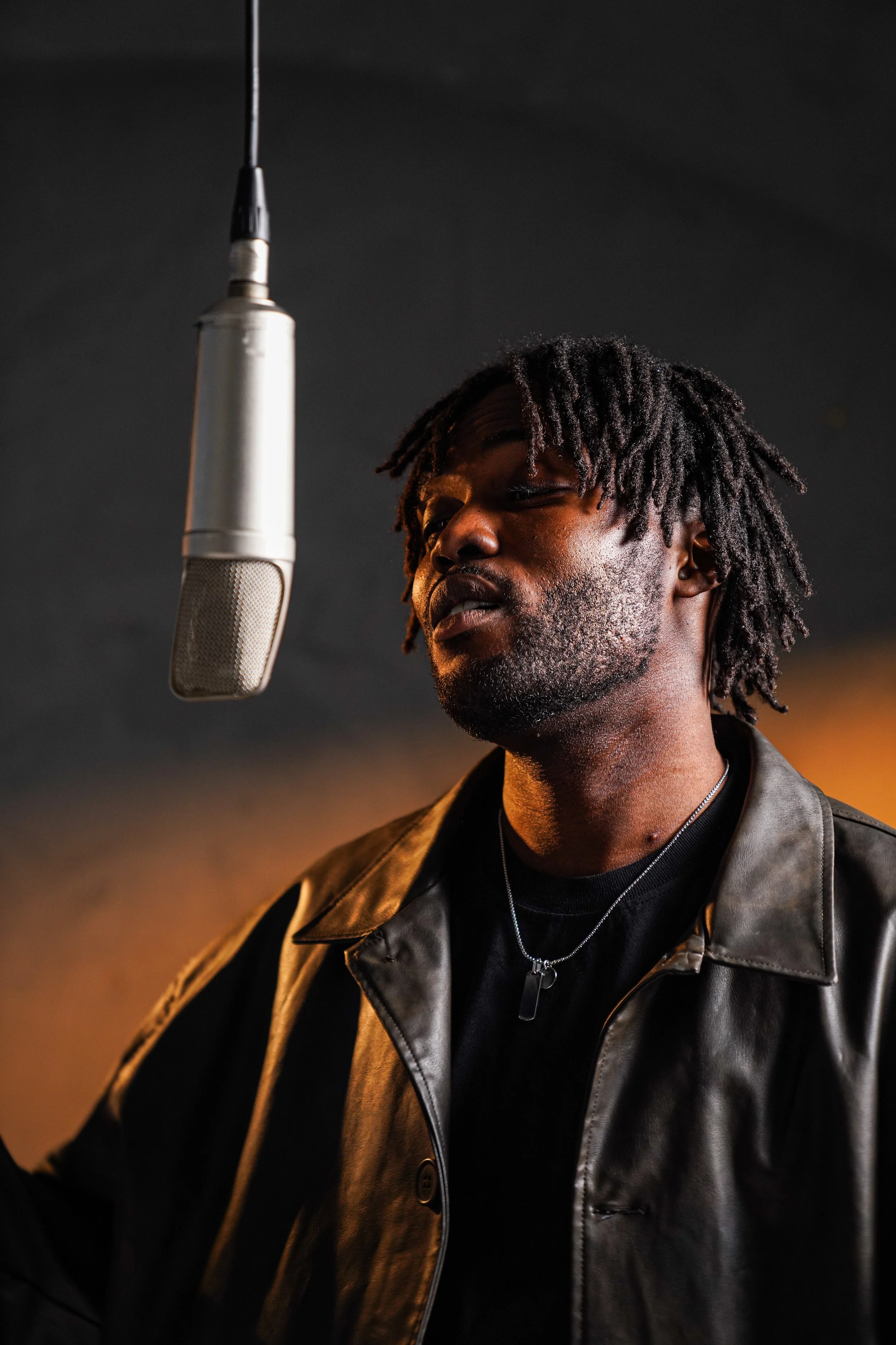 A man with dreadlocks singing or speaking into a microphone, wearing a black shirt and a leather jacket, in a recording studio.