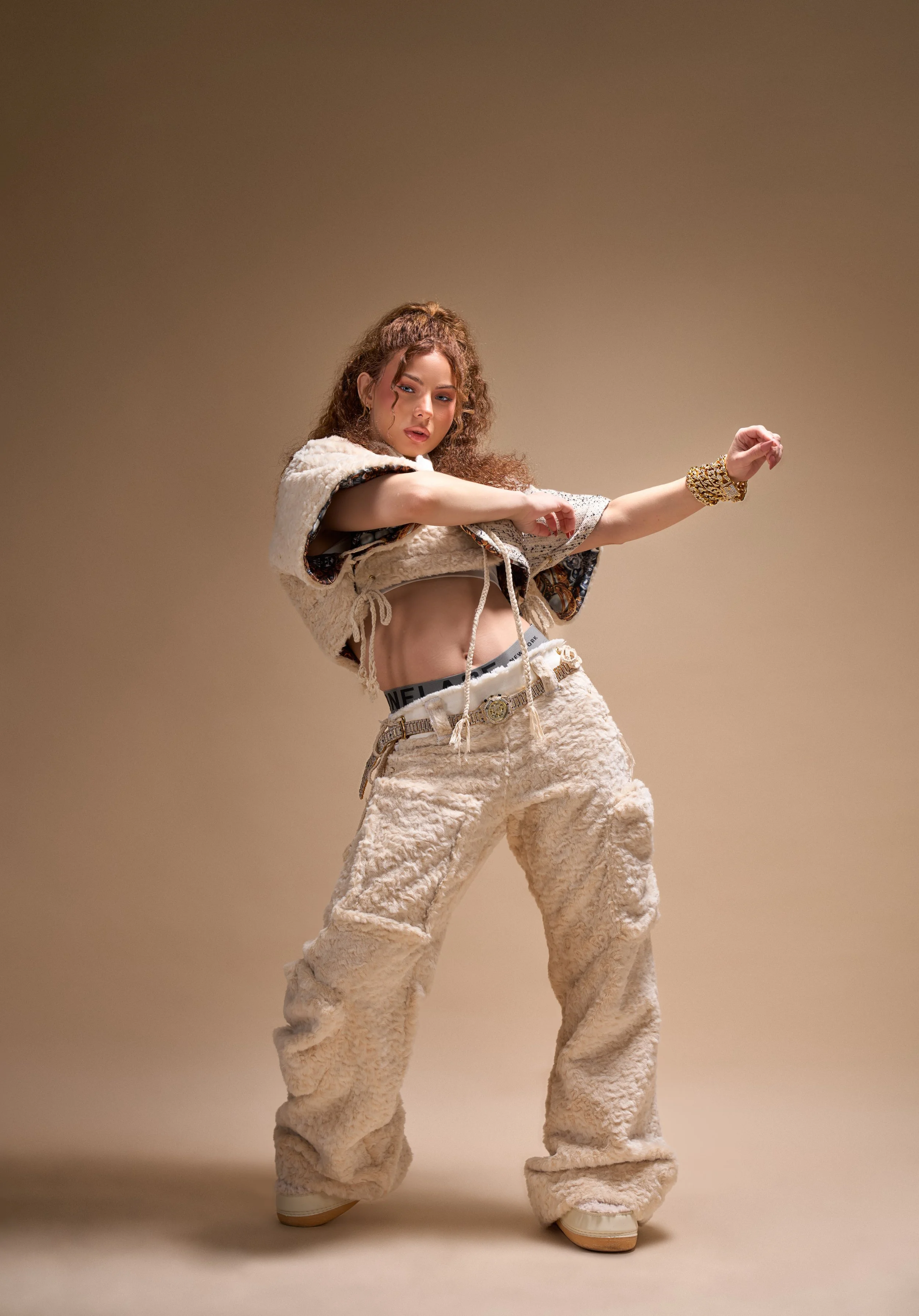 Young woman with curly hair dancing, wearing cream-colored plush pants, a cropped fur vest, and gold jewelry, against a neutral background.