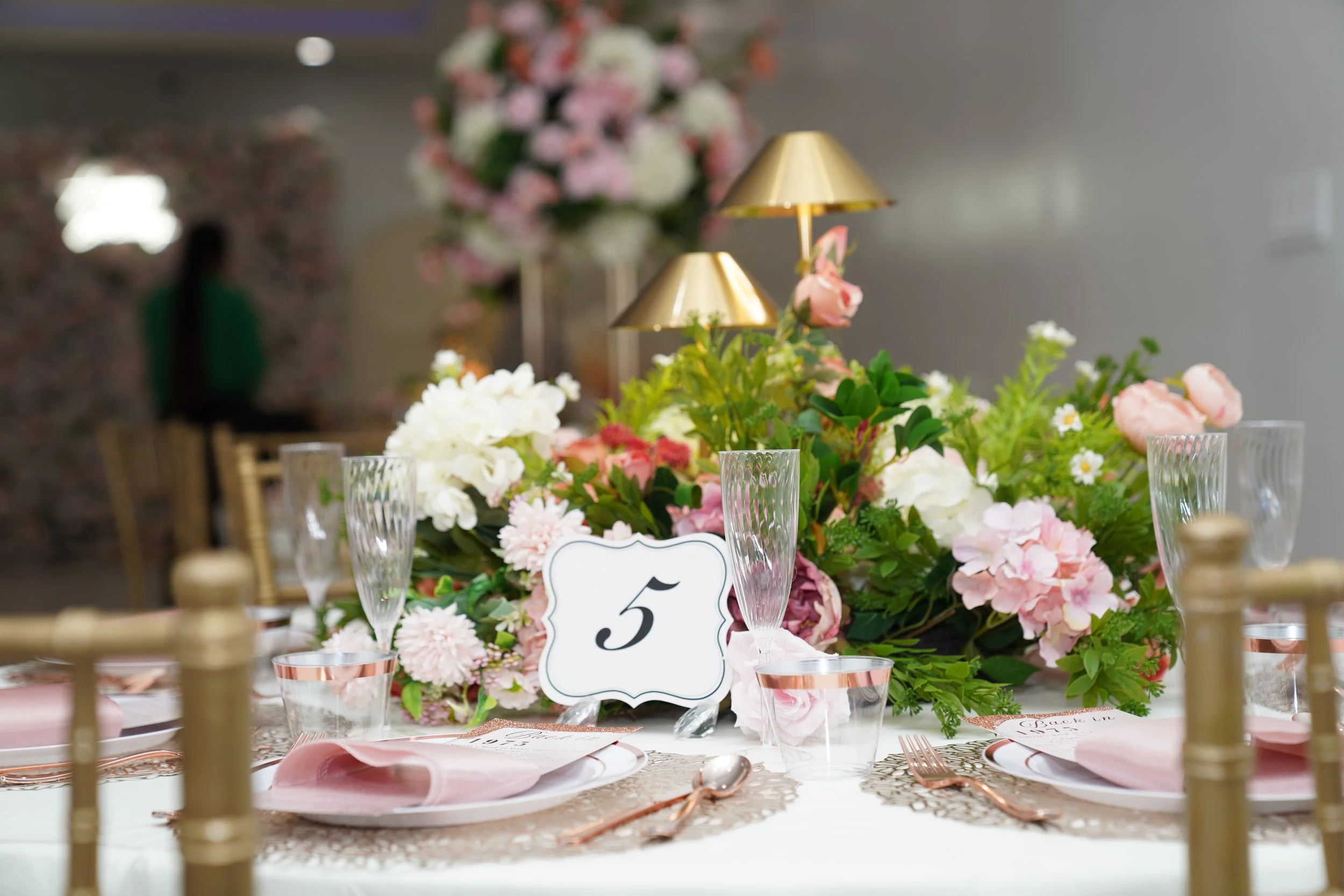 Elegant table setting with pink and white floral centerpiece, gold accents, glassware, plates, pink napkins, and a table number 5 card.