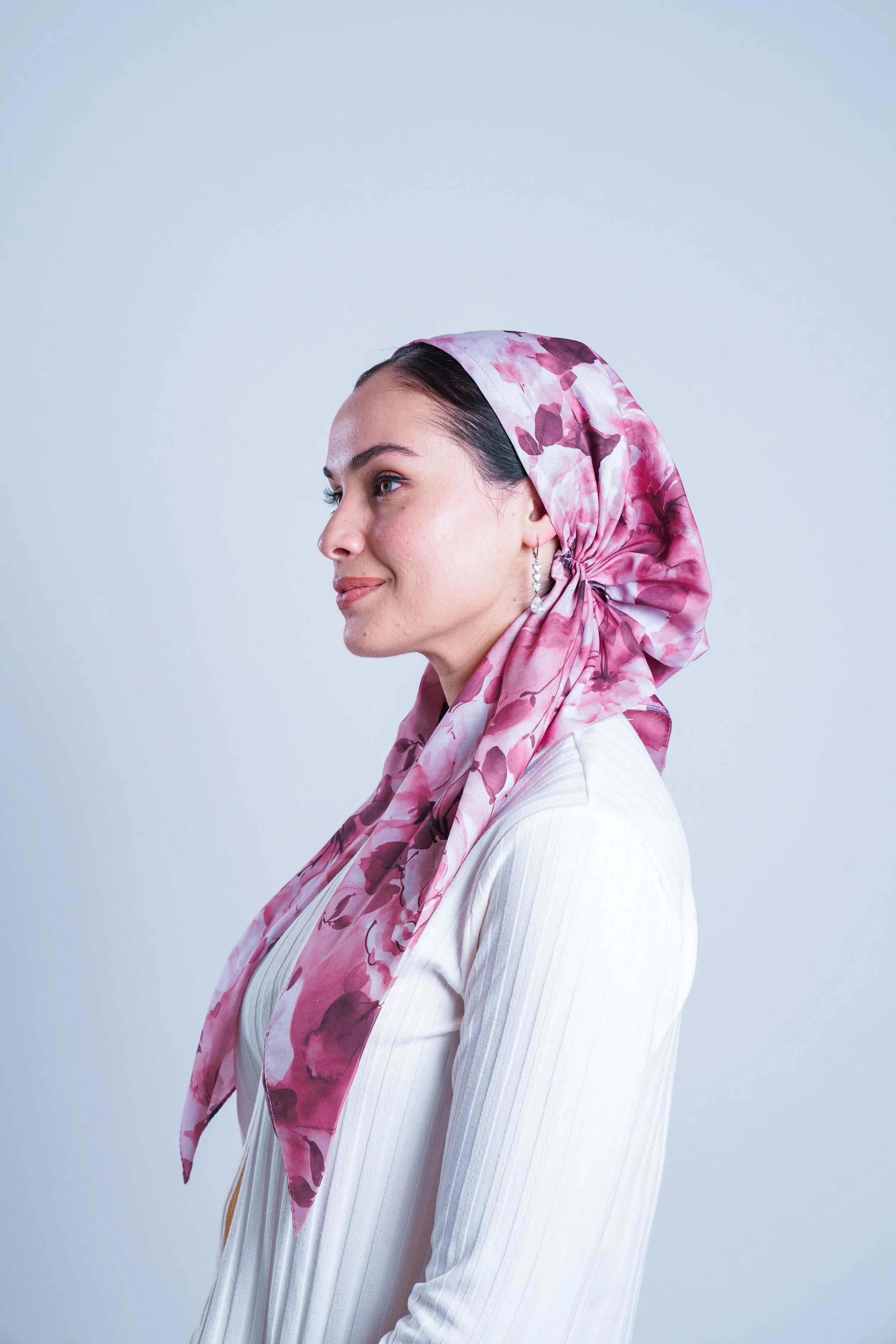 Profile of a woman with dark hair, wearing pearl earrings and a pink floral headscarf, dressed in a white striped shirt, against a plain light background.