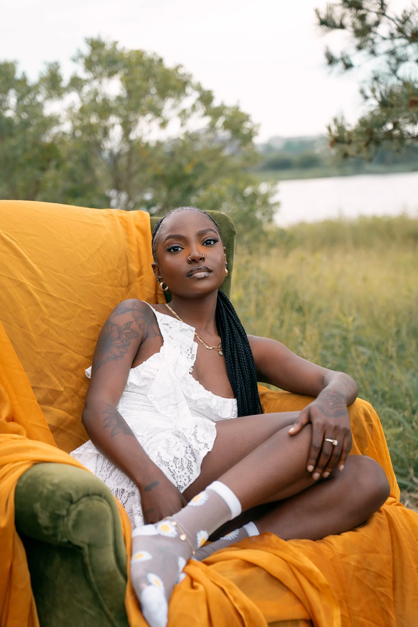 A young woman with dark skin and long braided hair sitting on a green armchair with an orange cover in an outdoor setting, surrounded by trees and grass, near a body of water during daytime, wearing a white lace dress and socks with daisies.
