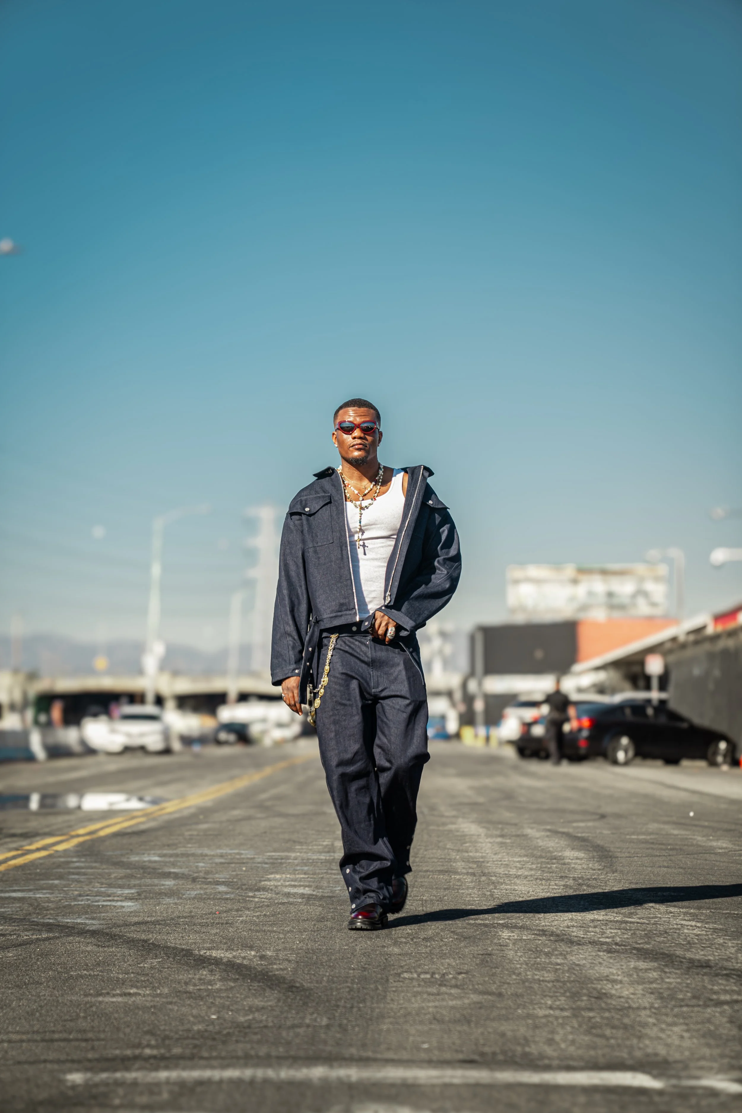 A man walking on a city street wearing dark denim jacket and pants, white shirt, sunglasses, and jewelry, with cars and buildings in the background under a clear blue sky.