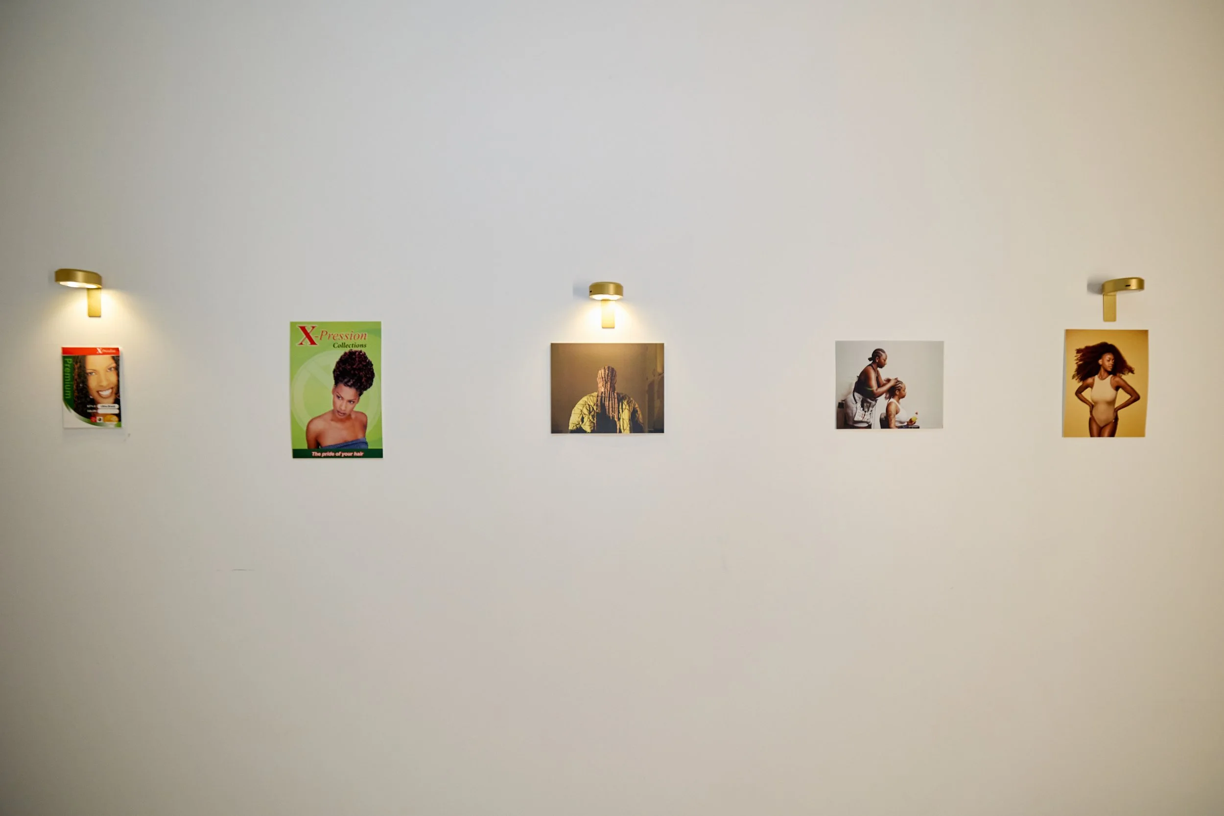 Gallery wall with five framed photographs and posters illuminated by small wall lights, showcasing diverse hairstyles and fashion, in an art or photo exhibition space.