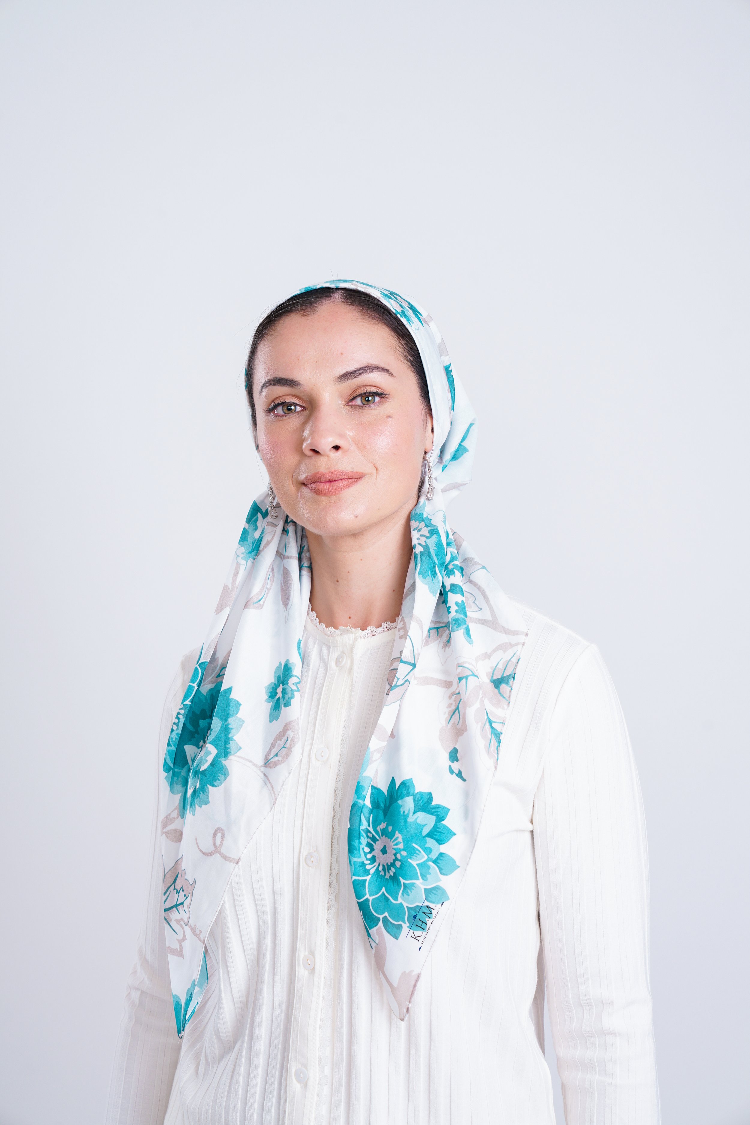 A woman with fair skin and dark hair wearing a white top and a white scarf with blue floral patterns, standing against a plain white background.