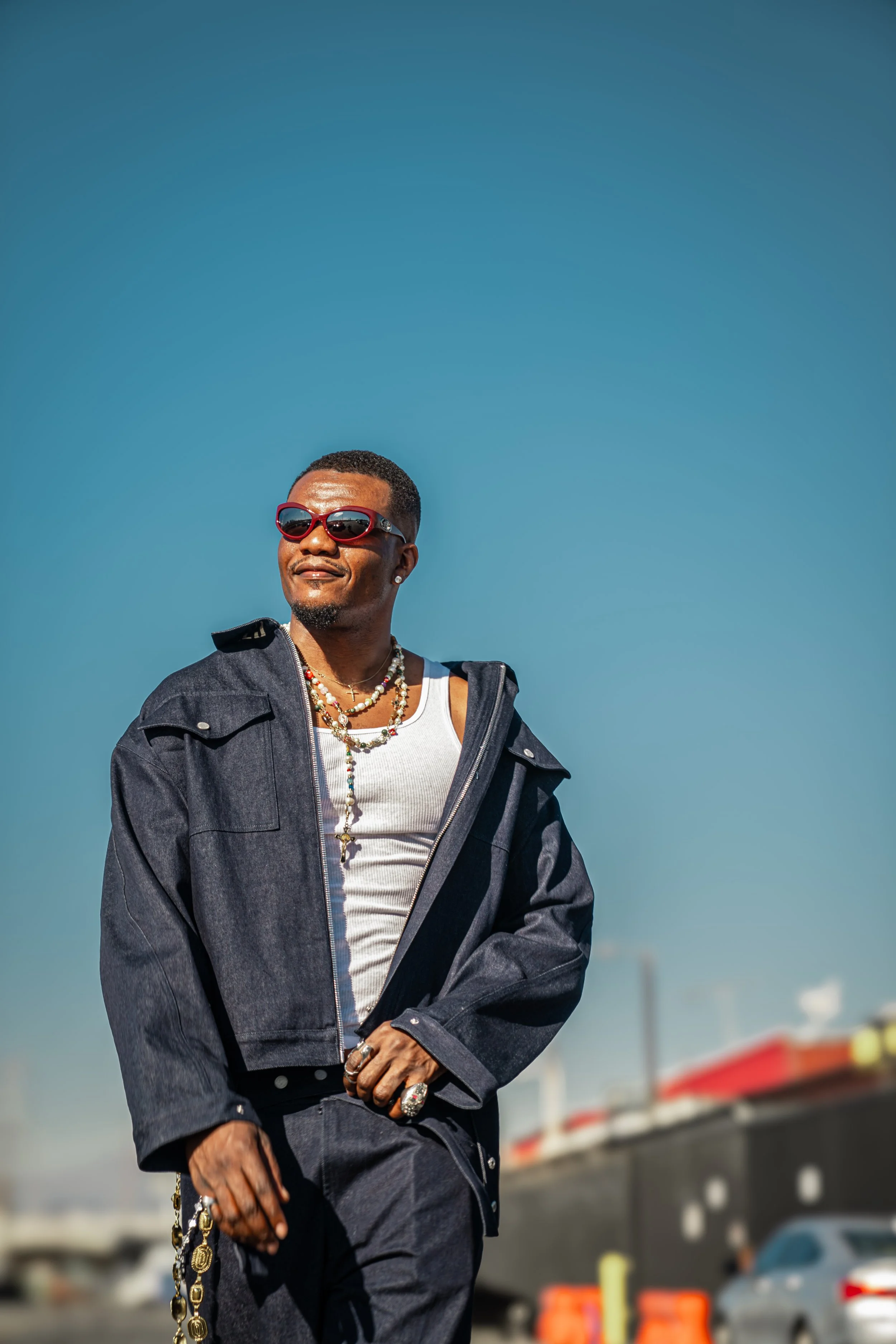 A man wearing sunglasses, a white tank top, a denim jacket, and jewelry, standing outdoors against a clear blue sky, with a commercial area in the background.