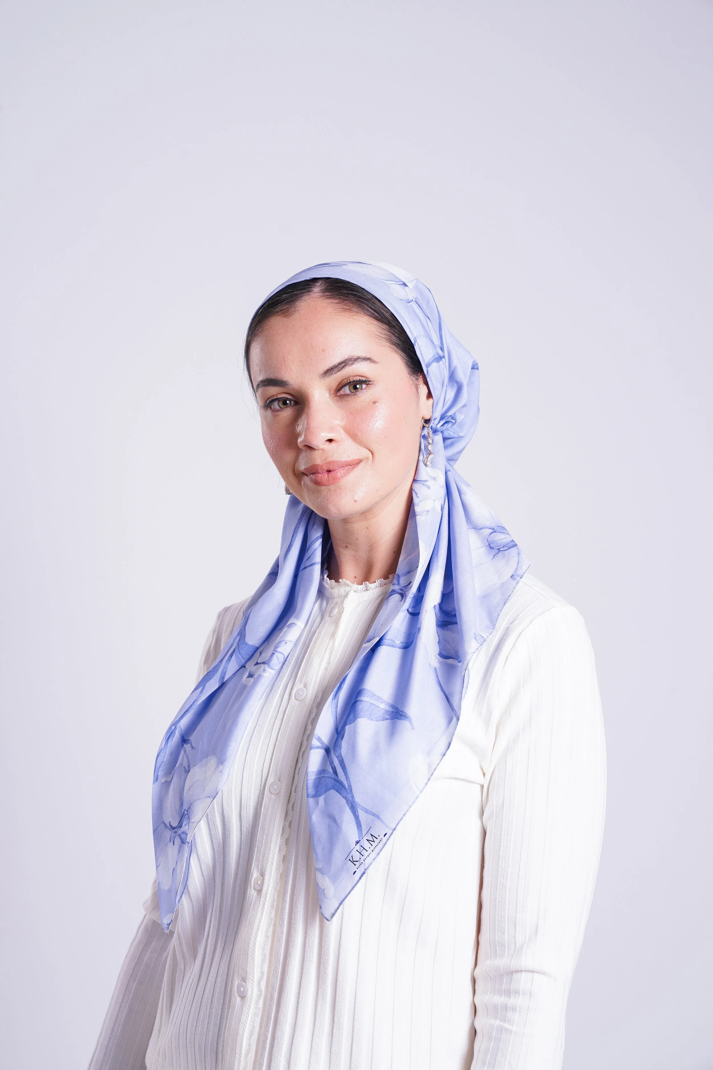A woman wearing a light blue silk headscarf and a cream-colored ribbed blouse, standing against a plain white background.