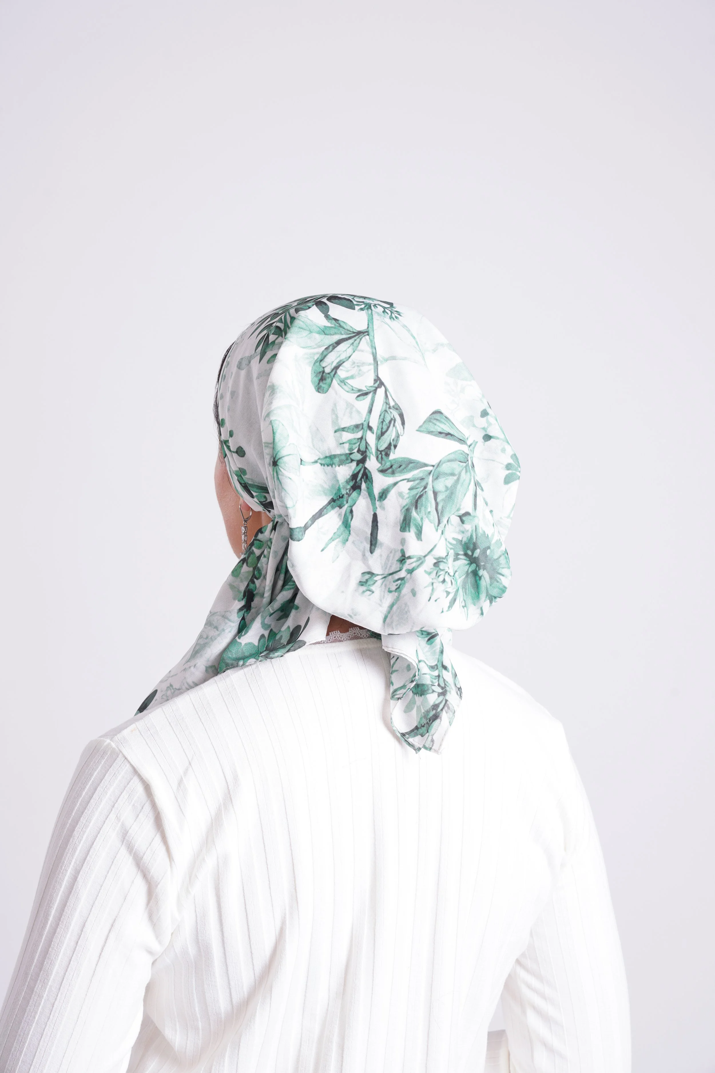 Back of a woman wearing a white top with a green floral patterned headscarf.