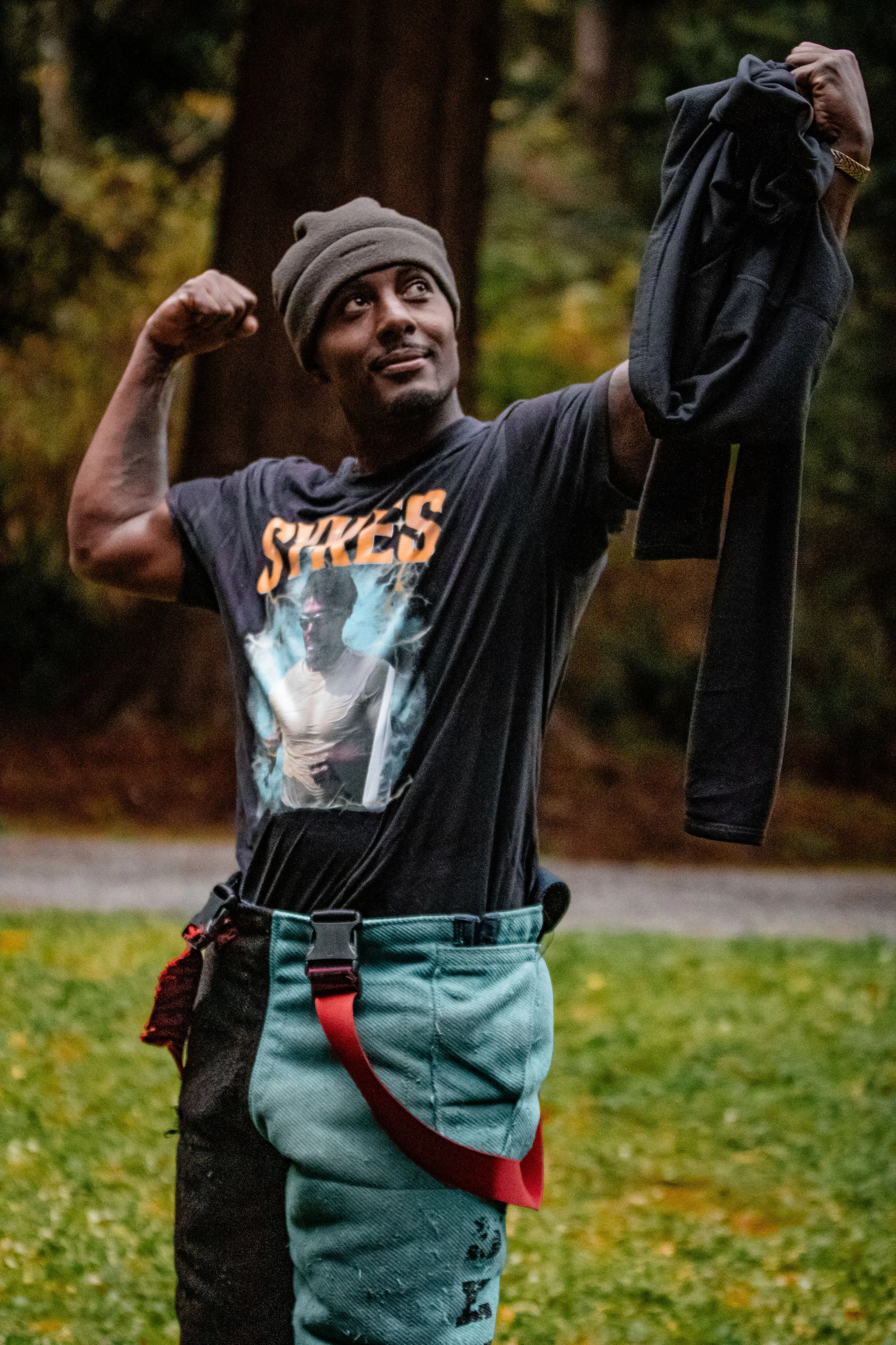 A man standing outdoors in a park, smiling and flexing his right arm, holding a jacket in his left hand. He is wearing a gray beanie, a graphic T-shirt, and two-tone pants with a red belt.