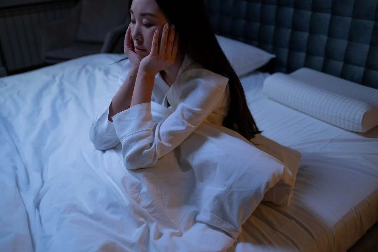 A woman sitting on a bed, looking thoughtful or worried, with her hands on her face, in a dimly lit bedroom.