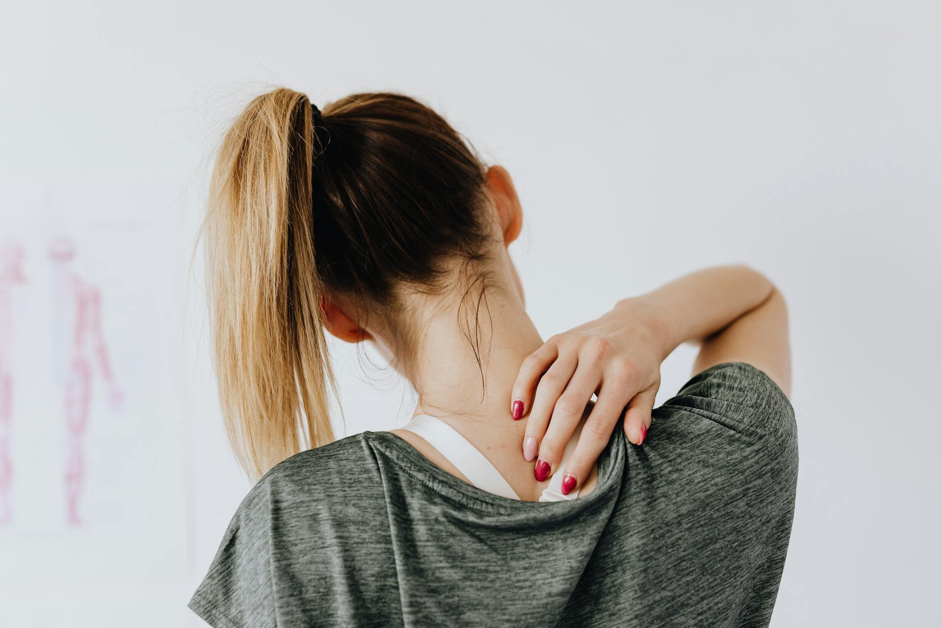 A woman with a ponytail and gray shirt reaches behind her neck to touch her shoulder, with a poster of human anatomy on the wall in the background.